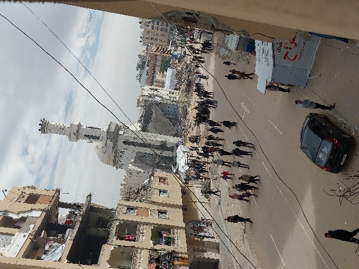 The image depicts a wide view of a street in Khan Yunis, Palestinian Territory, under an overcast sky, showing significant structural damage to buildings. In the center-background, a prominent white mosque with a minaret and dome stands partially damaged, surrounded by rubble. To the left, a multi-story building, likely residential, exhibits extensive destruction with missing walls, exposed interiors, and damaged balconies. Rubble is visible at its base.
The paved street running through the center is occupied by a large crowd of people, primarily adults and some children, walking in various directions, with many moving towards the background. A black hatchback car is parked on the right side of the street. Adjacent to the car, on the sidewalk, are several temporary tent-like structures or stalls, one with red Arabic text. Overhead, multiple black wires crisscross the scene. Further in the background, more urban structures are visible, some appearing damaged, and a large area of debris is present.