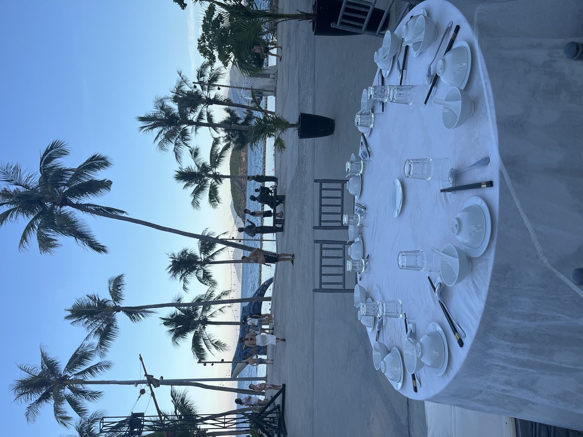 A circular table set for dining is in the foreground, covered with a white tablecloth and adorned with white bowls, plates, chopsticks, spoons, and glasses. In the midground, a paved area overlooks a body of water with a mountain range in the distance. Palm trees are scattered throughout the scene, some with large fronds, while others are smaller potted plants. Several people are visible in the midground, walking and conversing. A blue, whale-shaped sculpture is also present near the water's edge. The sky is clear and blue, suggesting it is either late afternoon or early morning.