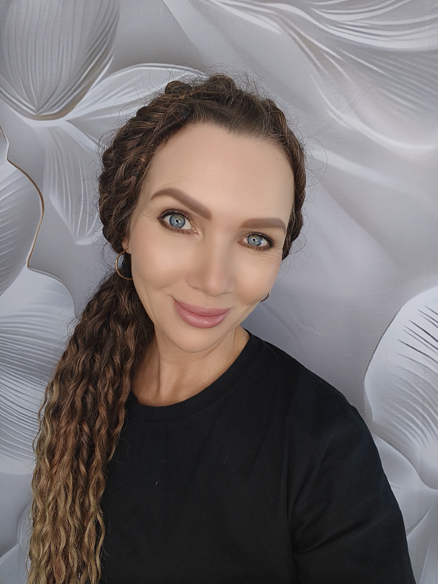 A woman with long, curly brown hair styled in braids and a ponytail, and striking blue eyes, smiles at the camera. She is wearing a black t-shirt and a gold hoop earring on her left ear. The background is a light gray wall with a white floral pattern. The woman is positioned in the center of the frame, with her head and shoulders visible.