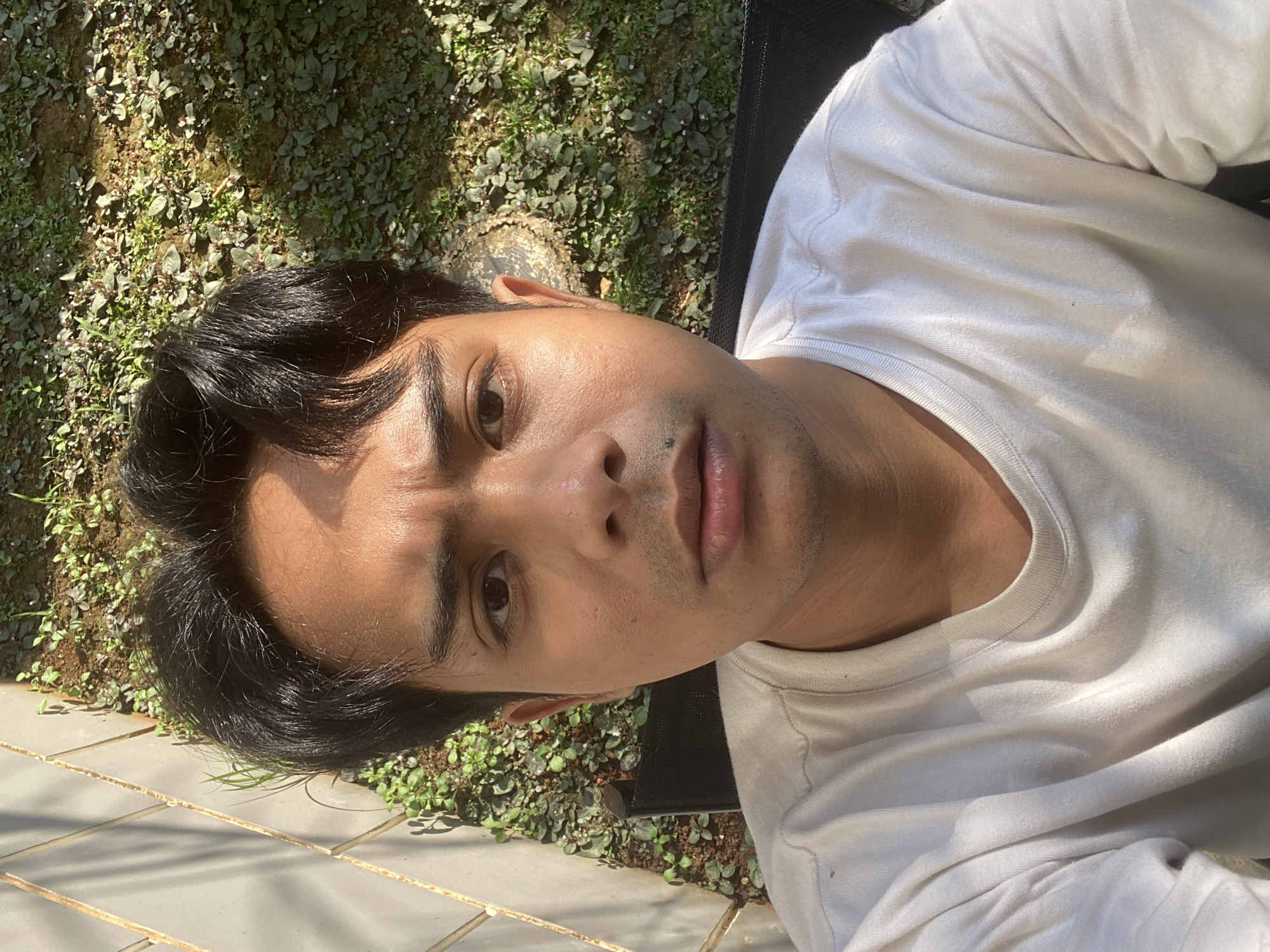 In Dukuh, Indonesia, a young man with dark hair and a light complexion is pictured in a close-up selfie. He is wearing a plain white t-shirt. The background shows a garden with green plants and soil, and to the left, a textured wall with some sunlight casting shadows. The man's expression is neutral, and he is looking directly at the camera.