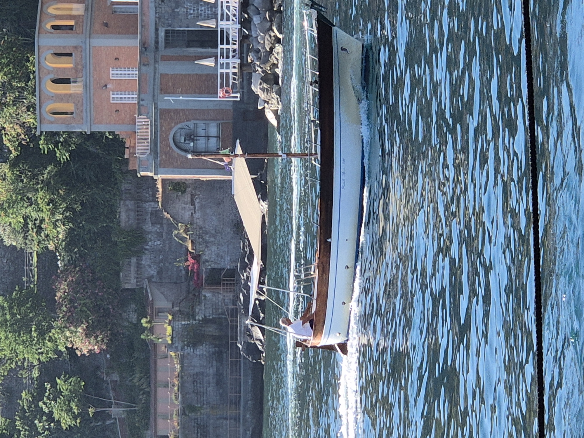 A man wearing a white t-shirt and dark shorts is operating a wooden motorboat on a body of water. The boat has a white hull with a wooden trim and a tan canopy providing shade. The boat is moving and creating a wake. In the background, there is a building with an arched facade and a tiered design. The building is constructed of brick and stone, and it has several windows and balconies. Beyond the building, there is lush green vegetation. The scene appears to be a sunny day.