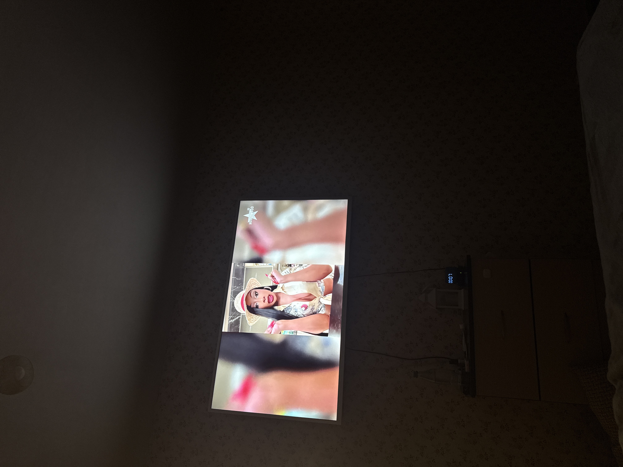 A television is mounted on a wall displaying an image of a woman wearing a straw hat and a light-colored outfit. The setting appears to be a room, with a patterned wall. A bedside cabinet with a digital clock is visible to the right of the television. The location is Mytishchi, Russia.