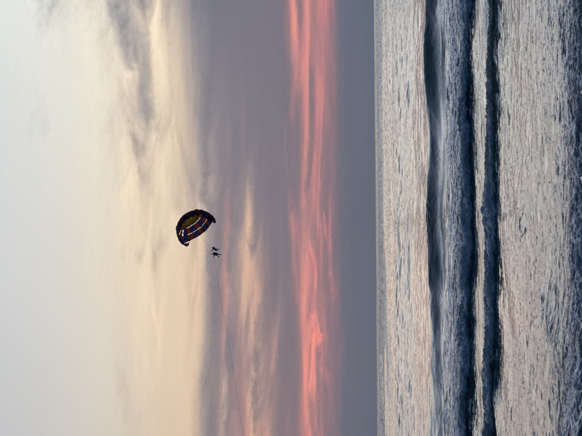 Two figures are parasailing above the ocean at dusk. They are attached to a colorful parasail, which is visible in the upper right quadrant of the image. The sky is a gradient of pale yellow and pink, with wispy clouds. The ocean below is calm, with gentle waves rolling towards the shore. The scene is likely on Patong Beach in Thailand, given the context provided.