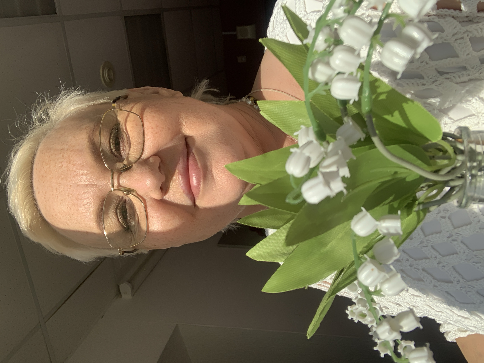 An eye-level close-up photograph shows a person with white hair and glasses, holding a bouquet of lily of the valley flowers. The person is indoors. The photograph was likely taken in Chaikovsky, Russia.