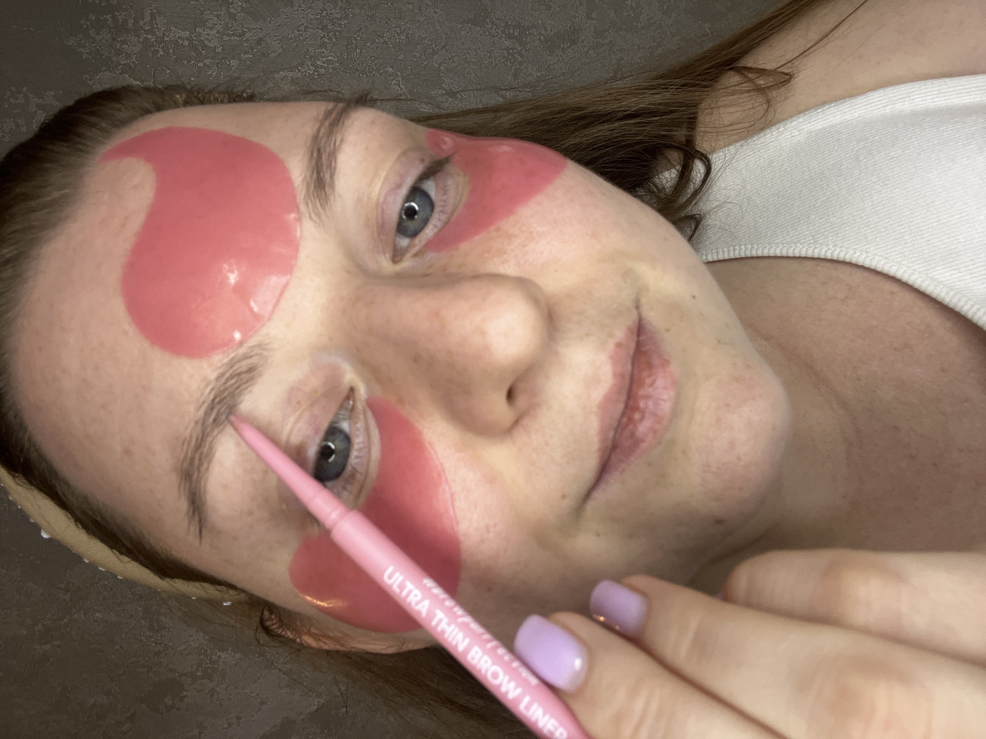 A woman is depicted indoors, possibly in a residential setting, applying eyebrow makeup. She has pink under-eye patches and a matching eyebrow pencil labelled "Ultra Thin Brow Liner." She has fair skin with freckles, blue eyes, and light brown hair. Her nails are painted a lilac color. She is wearing a white top. The location in the city of Раздолье, Russia, is not discernible from this image.