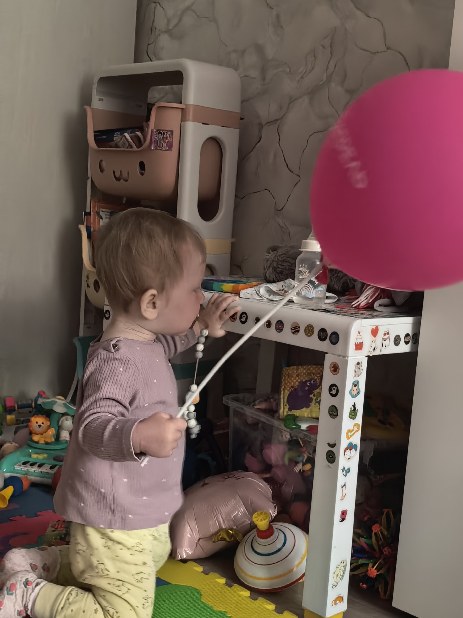 The image captures a toddler kneeling on colorful foam play mats in what appears to be an indoor play area, likely a home in Sterlitamak, Russia. The child, with light brown hair, is seen in profile, wearing a light purple long-sleeve shirt with subtle white dots and light yellow patterned pants. They are intently focused on a small white table or shelf unit, which is decorated with numerous cartoon stickers featuring various characters and shapes. The toddler holds a white stick adorned with various grey and white beads, engaging with something on the table or simply exploring the toy.
On the table, a baby bottle with liquid and some fabric items are visible. In the upper right corner, a large, bright pink balloon is partially seen, bearing faint white text that is not clearly decipherable but suggests a celebratory message.
Behind the child and to the left, a multi-tiered toy organizer or shelf unit, in white and pink, houses a cute pink bin with a carved face, holding various items. The background wall features a distinctive grey and white textured pattern. The floor is covered with interlocking green, yellow, and red foam mats. Various toys are scattered around the floor, including a green toy piano with a lion figure, a large spinning top, a deflated pink foil balloon, and a clear bin filled with other toys. The scene depicts a quiet moment of a child's focused play and exploration within a cluttered yet cozy domestic environment. No specific time of day or weather is discernible.