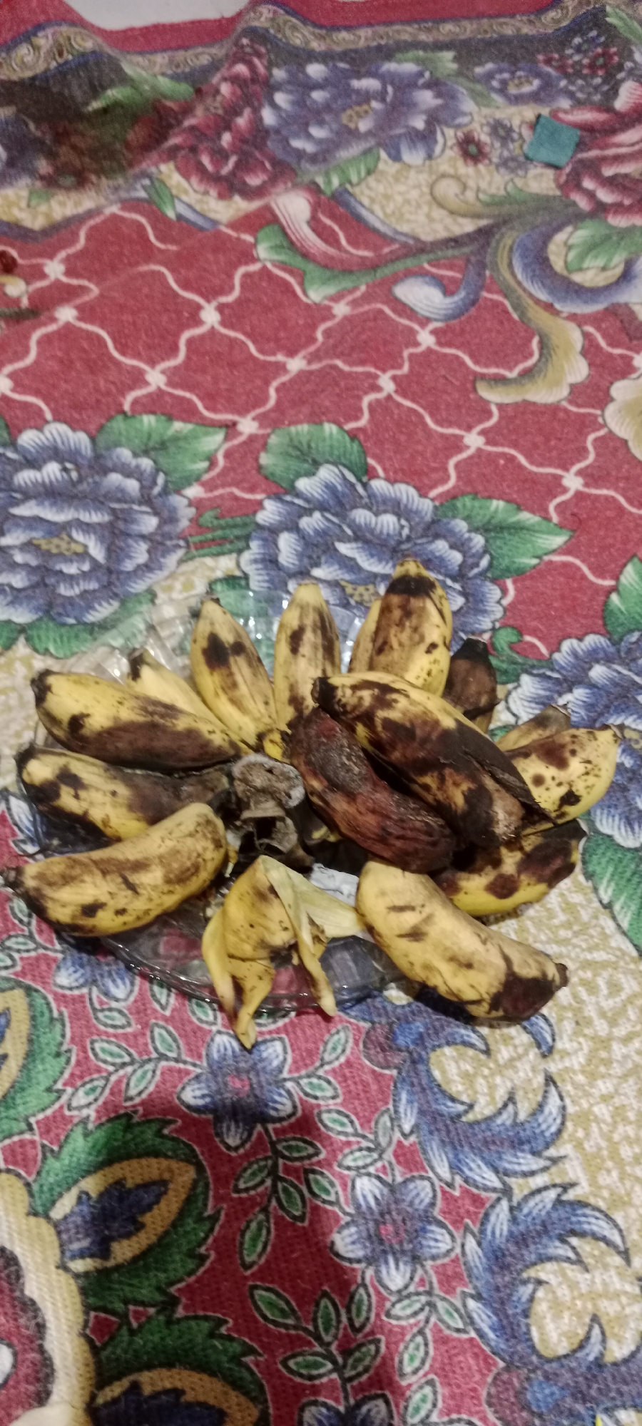 A plate of overripe bananas sits on a patterned tablecloth. The bananas are yellow with significant brown and black spotting, indicating they are past their prime. Some of the peels are partially removed, revealing the darkened fruit beneath. The tablecloth features a repeating floral pattern with red, blue, green, and yellow colors. The lighting is dim, and the image appears to be taken from a slightly elevated angle. The scene is indoors, on a flat surface.