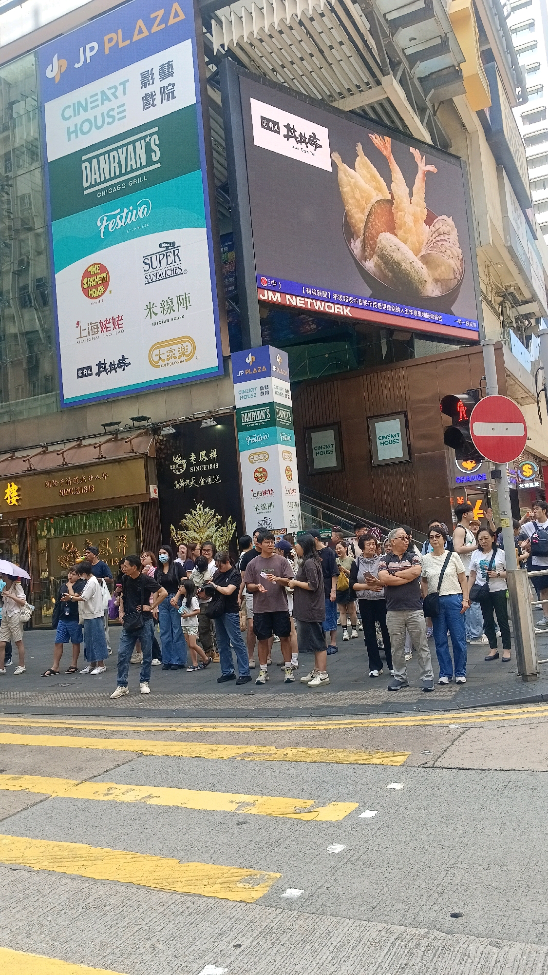 This image captures a street scene in Hong Kong, China, focusing on a pedestrian crossing and the surrounding commercial buildings. The foreground shows a yellow and grey pedestrian crossing on an asphalt road. In the background, a crowd of people are standing on the sidewalk, some appearing to wait for a traffic signal.
Several large electronic billboards are prominent on the buildings. One billboard displays the "JP PLAZA" logo and various commercial advertisements, including "CINEART HOUSE," "DANRYAN'S CHICAGO GRILL," and "Festiva." Another large screen shows an advertisement for "Don Tei" restaurant, featuring a bowl of tempura and rice. Text in both English and Chinese is visible on the billboards and building signage, indicating commercial activity.
The architecture suggests a busy urban environment with multi-story buildings. Traffic lights and road signs are also visible, including a red circular sign with a white horizontal bar, indicating a prohibition, and a red pedestrian signal showing a standing figure. The overall impression is of a bustling commercial district in Hong Kong, with people navigating the urban landscape and engaging with digital advertising.