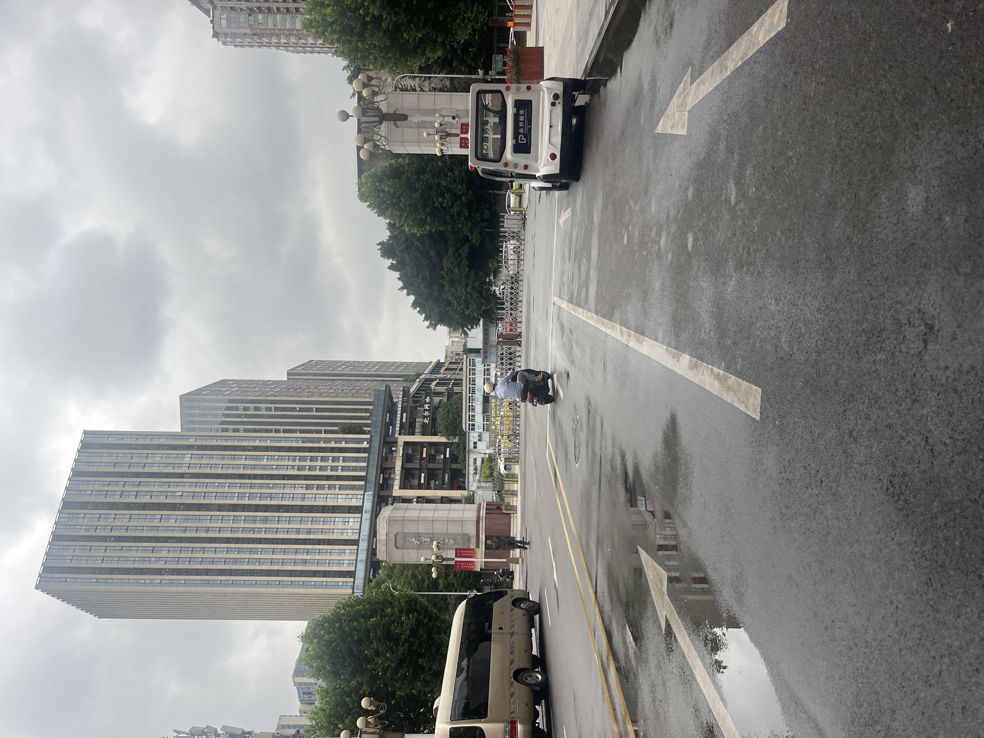 A wet, overcast day in Yuzhong District, China, where a wide asphalt road with directional arrows is seen reflecting the muted sky and surrounding buildings. On the right side of the road, a small white electric vehicle with "永升服务" (Yong Sheng Fu Wu) printed on its rear is parked. Further down the road, a person rides a black motorcycle. To the left, a gold minibus is stopped. In the background, a large, modern building with many windows dominates the skyline. Trees and decorative streetlights line the street.