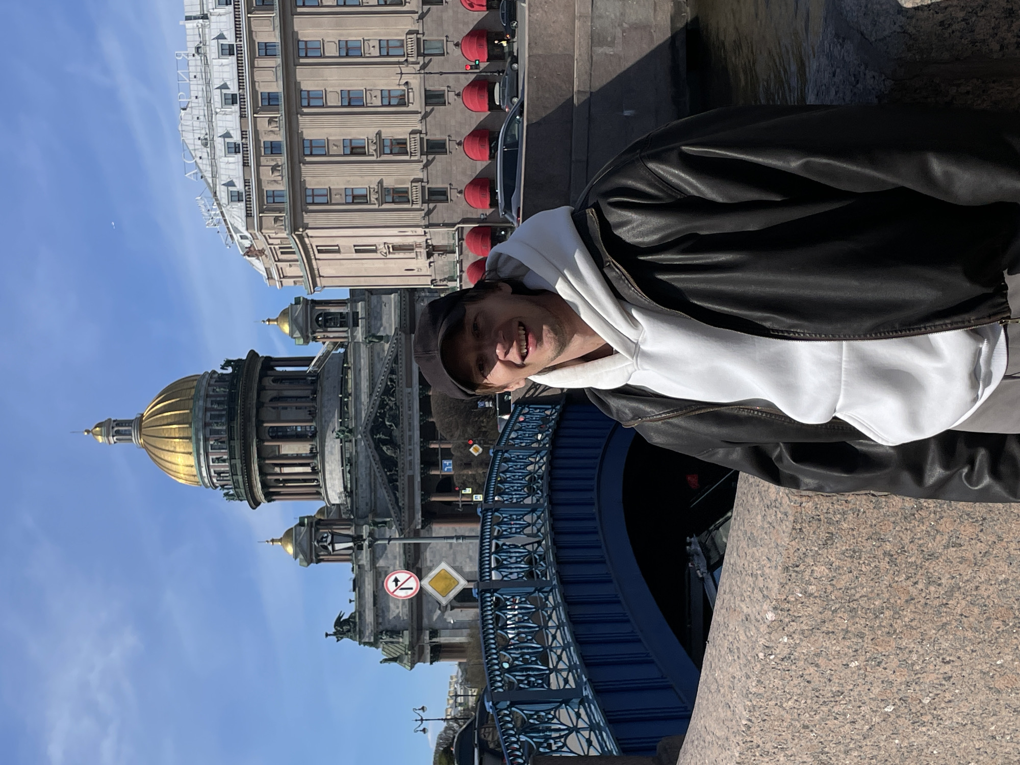 A young man poses smiling for a photo in Saint Petersburg, Russia, against a backdrop of iconic architecture. He is positioned on the right side of the frame, from the chest up, wearing a white hoodie, a black leather jacket, and a dark baseball cap. He looks directly at the camera with a wide smile.
The scene takes place outdoors on a bright, sunny day, with a clear blue sky. Behind the man and slightly to his left stands St. Isaac's Cathedral, clearly identifiable by its magnificent golden dome and neoclassical design. Further back, another large building with numerous windows and several red dome-shaped awnings is visible. A dark-colored car is partially seen parked near this building.
In the mid-ground, a blue ornate bridge railing stretches across the image. The man appears to be standing on a granite-like stone parapet in the foreground.
Visible text includes two road signs to the left of the man and above the bridge: a circular sign with a red border and a black arrow depicting a U-turn (likely indicating "no U-turn" or "no entry" in that direction) and a yellow diamond-shaped sign next to it, which is a common priority road sign. On the roofline of the building behind the man, some Cyrillic letters are visible, appearing to spell "БАНКЪ" (Bank). The strong sunlight creates clear shadows, indicating it is late morning or early afternoon.