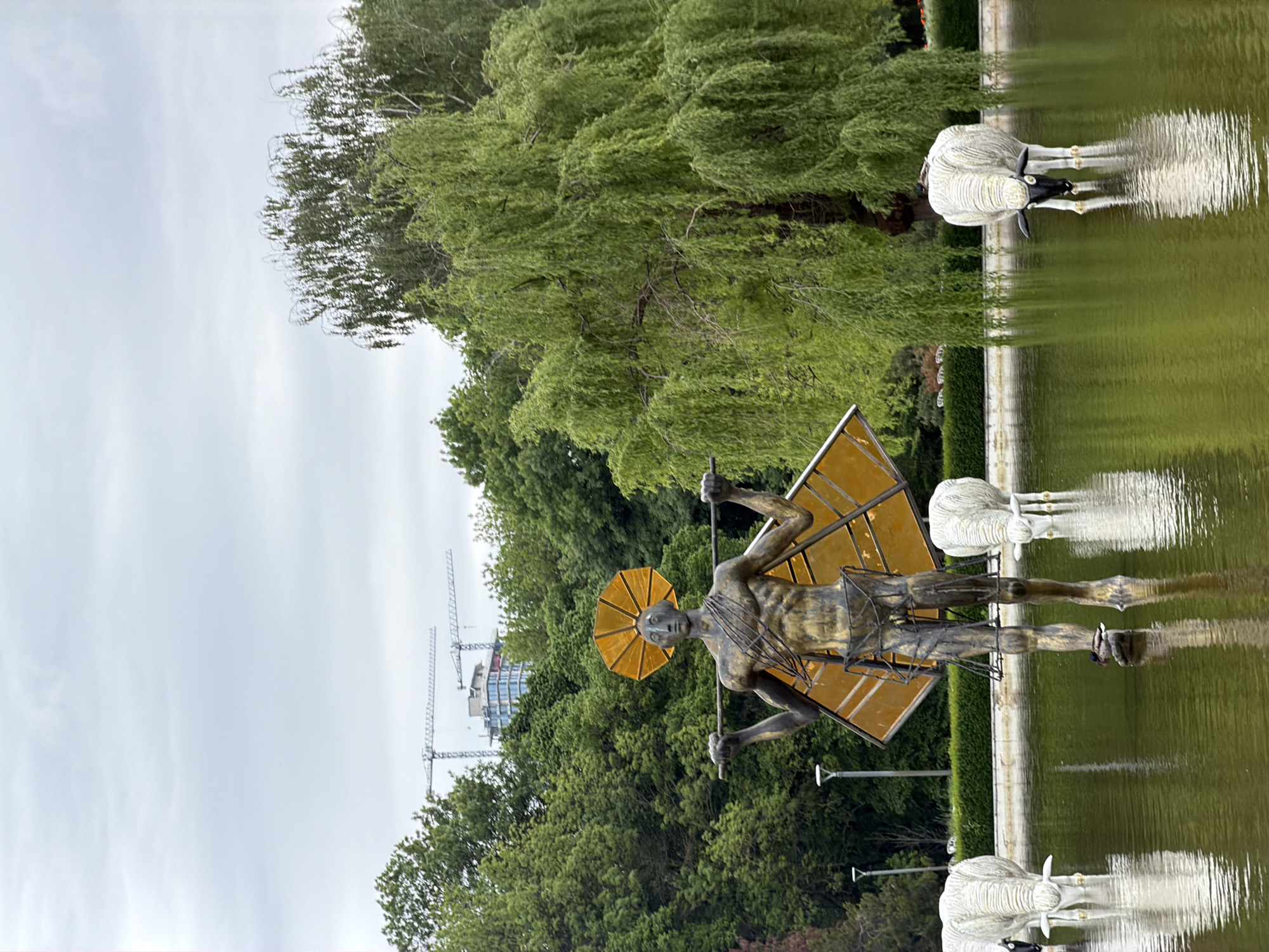 The image shows a statue of a man with a yellow umbrella-like halo and a yellow angular frame, standing in a pond with a reflection of himself and three sheep-like sculptures drinking from the water. Green trees are behind the statue, and in the distance, there are buildings and construction cranes. The location is the park in Sofia, Bulgaria.