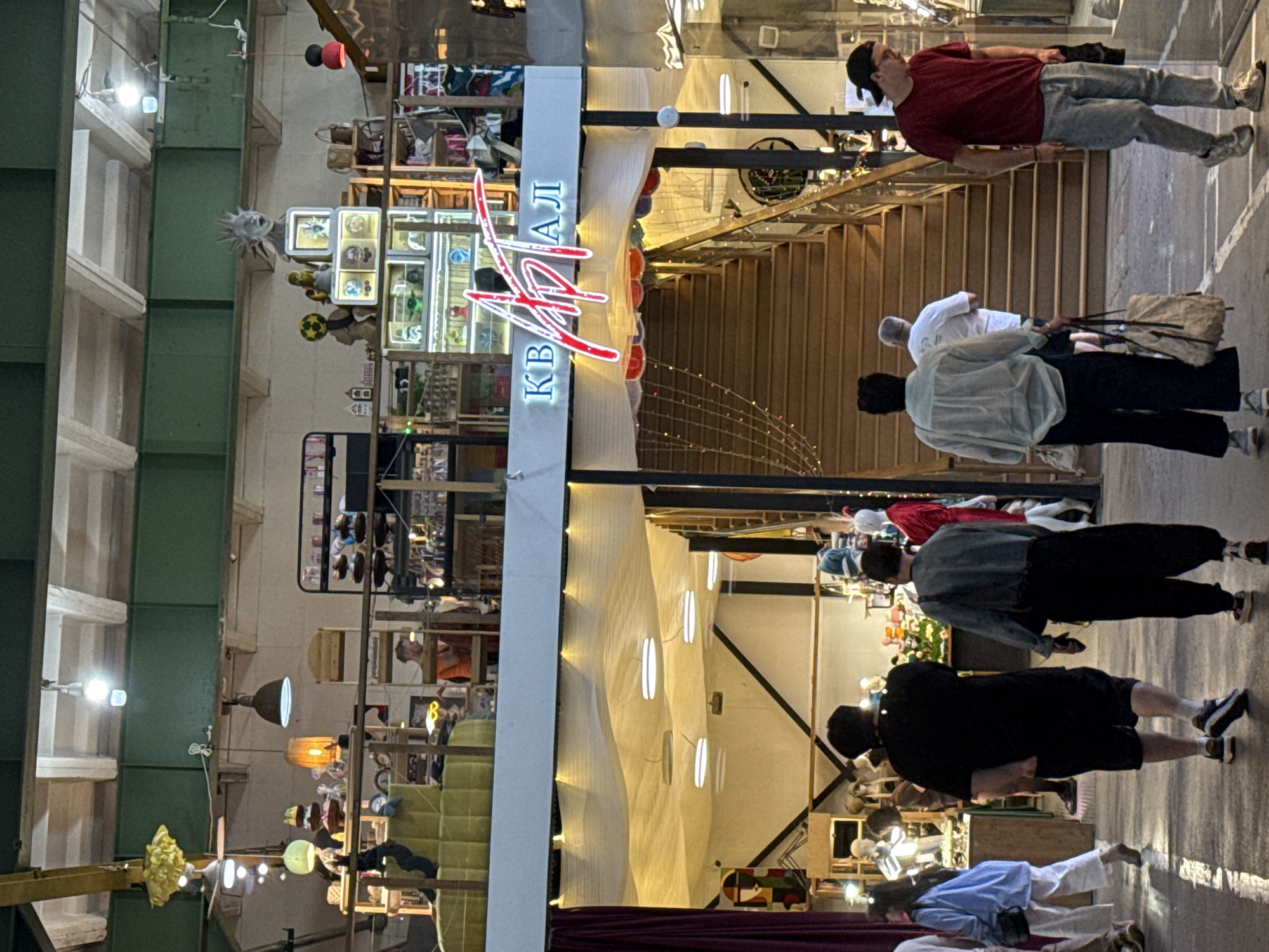 The image depicts the interior of the "KVARTAL" (КВАРТАЛ) market in Saint Petersburg, Russia, a multi-level indoor market hall. In the foreground, several individuals are walking across a polished floor marked with white lines. To the left, a person in a blue shirt and white pants walks away from the camera. In the center, a person in a black t-shirt and shorts walks away, followed by a person in a light blue denim jacket and dark pants carrying a large tote bag. To the right, a person in a red t-shirt and light blue jeans walks towards a display case.
The market has an industrial aesthetic with exposed beams and pipes in the ceiling. The middle level features open retail spaces with shelves displaying various items, including what appear to be decorative objects and clothing. A prominent red neon sign with Cyrillic lettering, "KVARTAL," is visible above a set of wooden stairs that lead up to the upper levels. The stairs are decorated with strings of small, colorful lights. The upper levels show more retail displays and lighting fixtures, including chandeliers and spotlights. The overall atmosphere suggests a vibrant and bustling marketplace.
