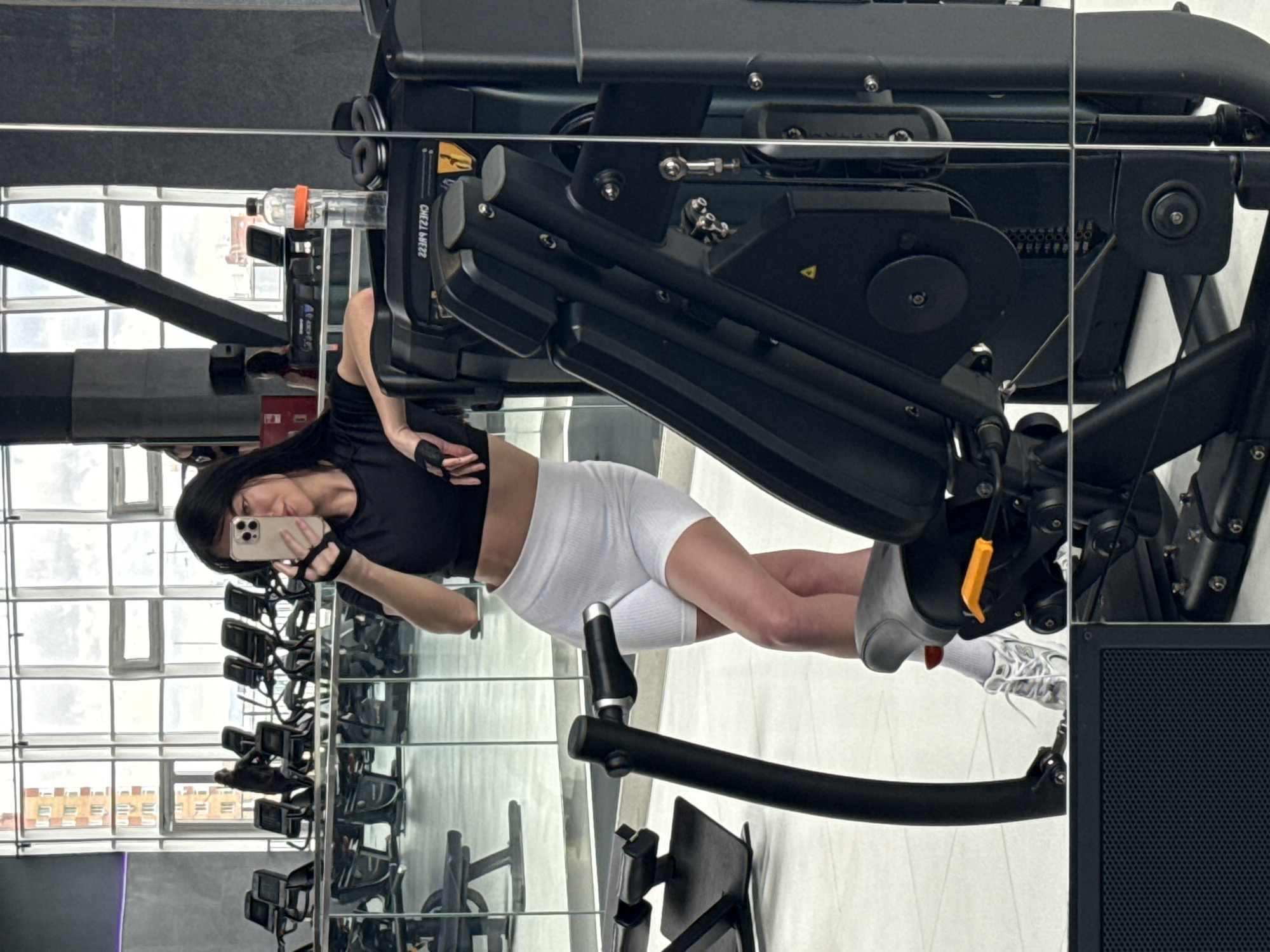 A mirror image captures a woman at a gym in Krasnoyarsk, Russia. She holds a smartphone, presumably taking a selfie. She is wearing a black sports bra, white shorts, socks, and gym gloves. The gym contains various weight machines, with windows revealing a cityscape outside.