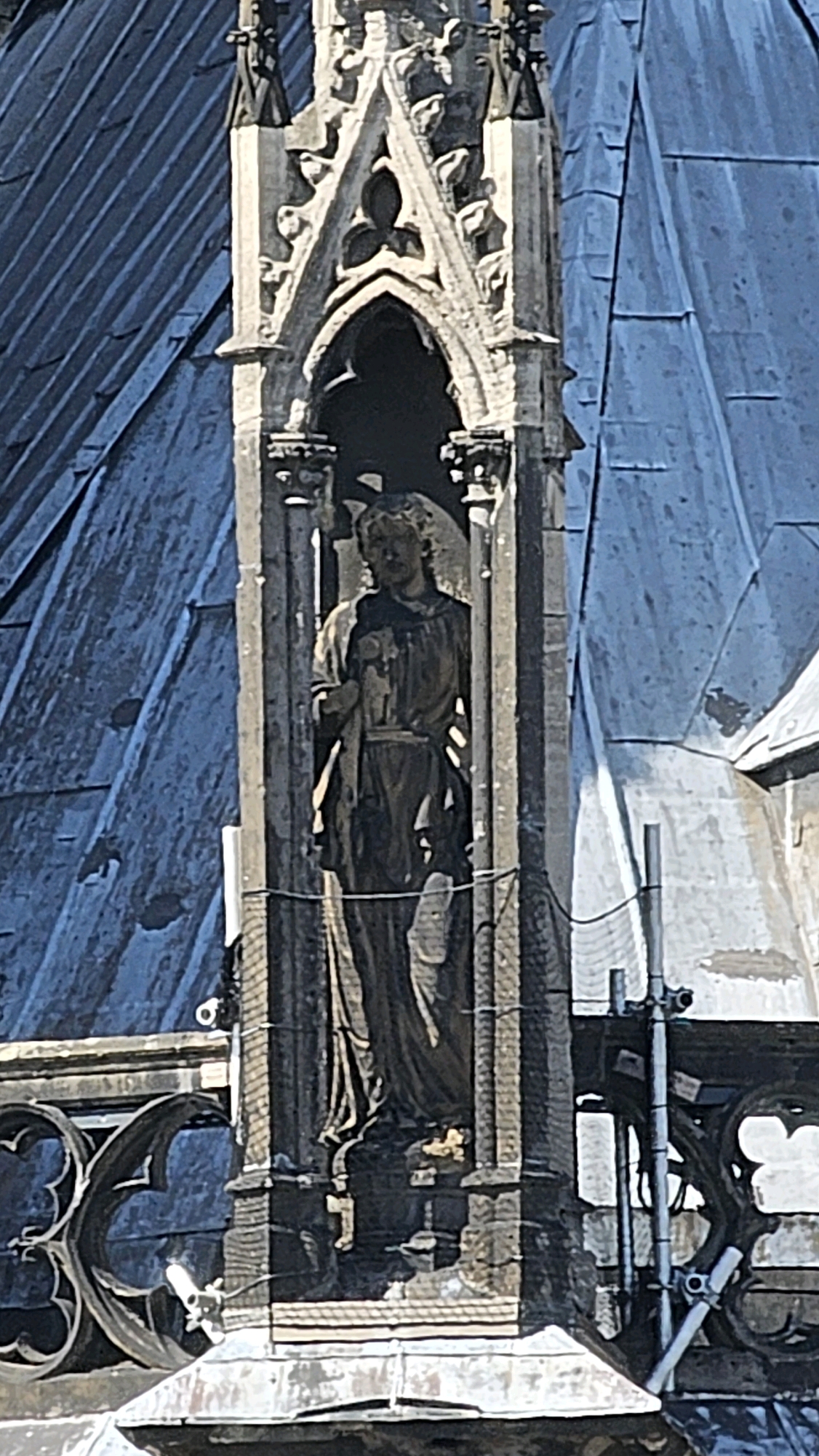 This image shows a detailed close-up of a stone statue of a robed figure within a gothic architectural niche on the exterior of a building. The statue is positioned in a vertical recessed section of the building's facade, framed by ornate stone carvings. The figure appears to be holding an object in its left hand, possibly a book or scroll. The statue's facial features are somewhat indistinct due to wear and lighting.
The architectural elements suggest a historical religious building. The statue is surrounded by detailed stonework, including pointed arches and decorative moldings, typical of Gothic architecture. Behind the statue, parts of a blue-tiled roof are visible, angled and textured, indicating the building's structure. Some scaffolding and support poles are also visible to the right of the statue, suggesting ongoing maintenance or restoration work on the building.
The statue and its niche are part of a larger architectural structure, likely a cathedral or church, given the style and ornamentation. The building is situated in a city environment, as evidenced by the construction and the scale of the architecture. The overall scene conveys a sense of historical significance and architectural grandeur. This specific statue is located on the exterior of Cologne Cathedral (Kölner Dom) in Cologne, Germany, specifically on one of its numerous spires or tower elements.
