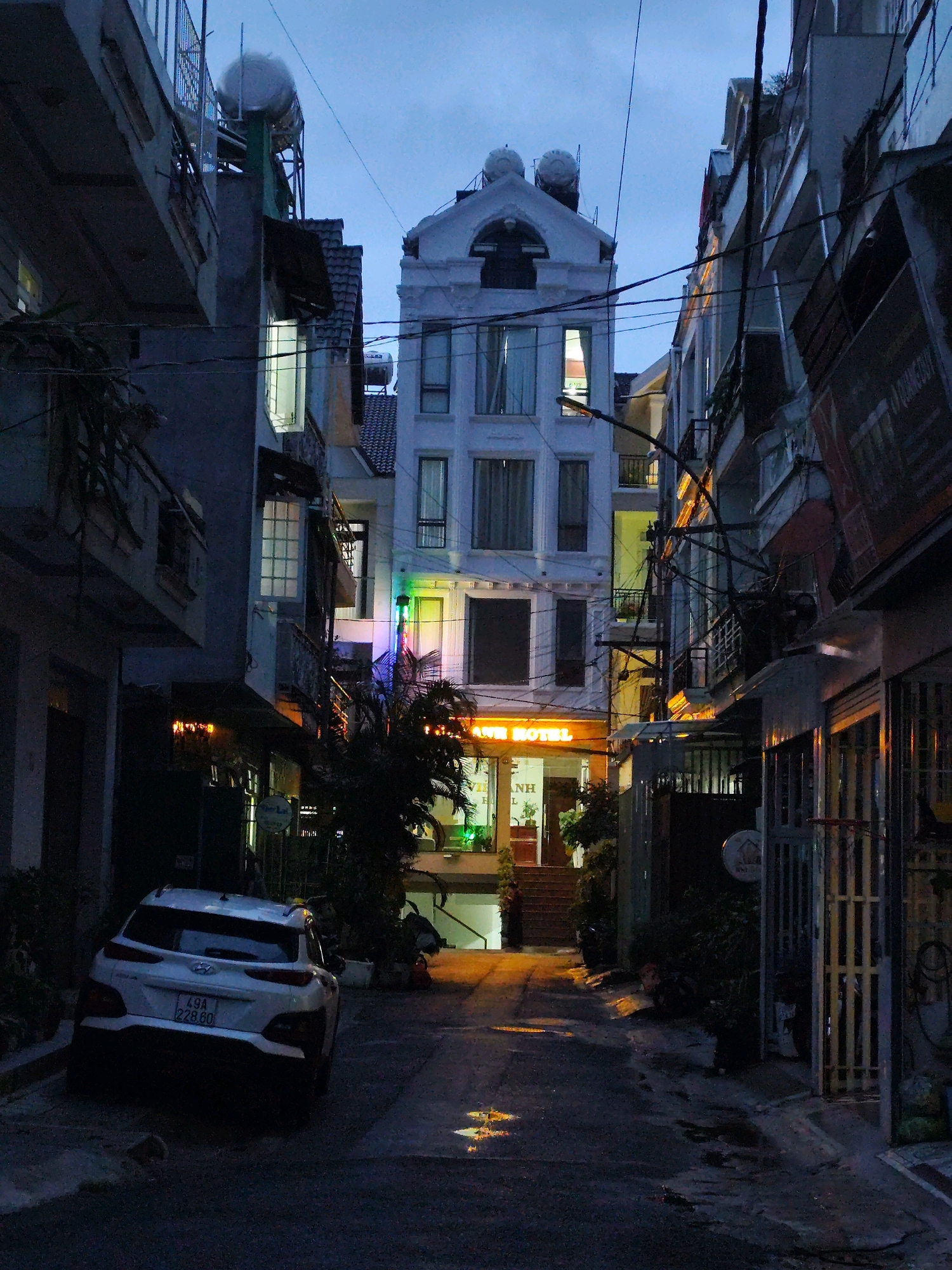 The image depicts a narrow street in Da Lat, Vietnam, at dusk. The street is lined with multi-story buildings, some appearing residential and others commercial, with the most prominent being "VINH ANH HOTEL" which is illuminated by orange neon lights. Electrical wires crisscross the sky, and several water tanks are visible on the rooftops. A white car with a license plate reading "49A 228.60" is parked on the left side of the street. The ground is wet, with reflections indicating recent rain. The atmosphere is dim and slightly moody, typical of an urban evening in Southeast Asia.