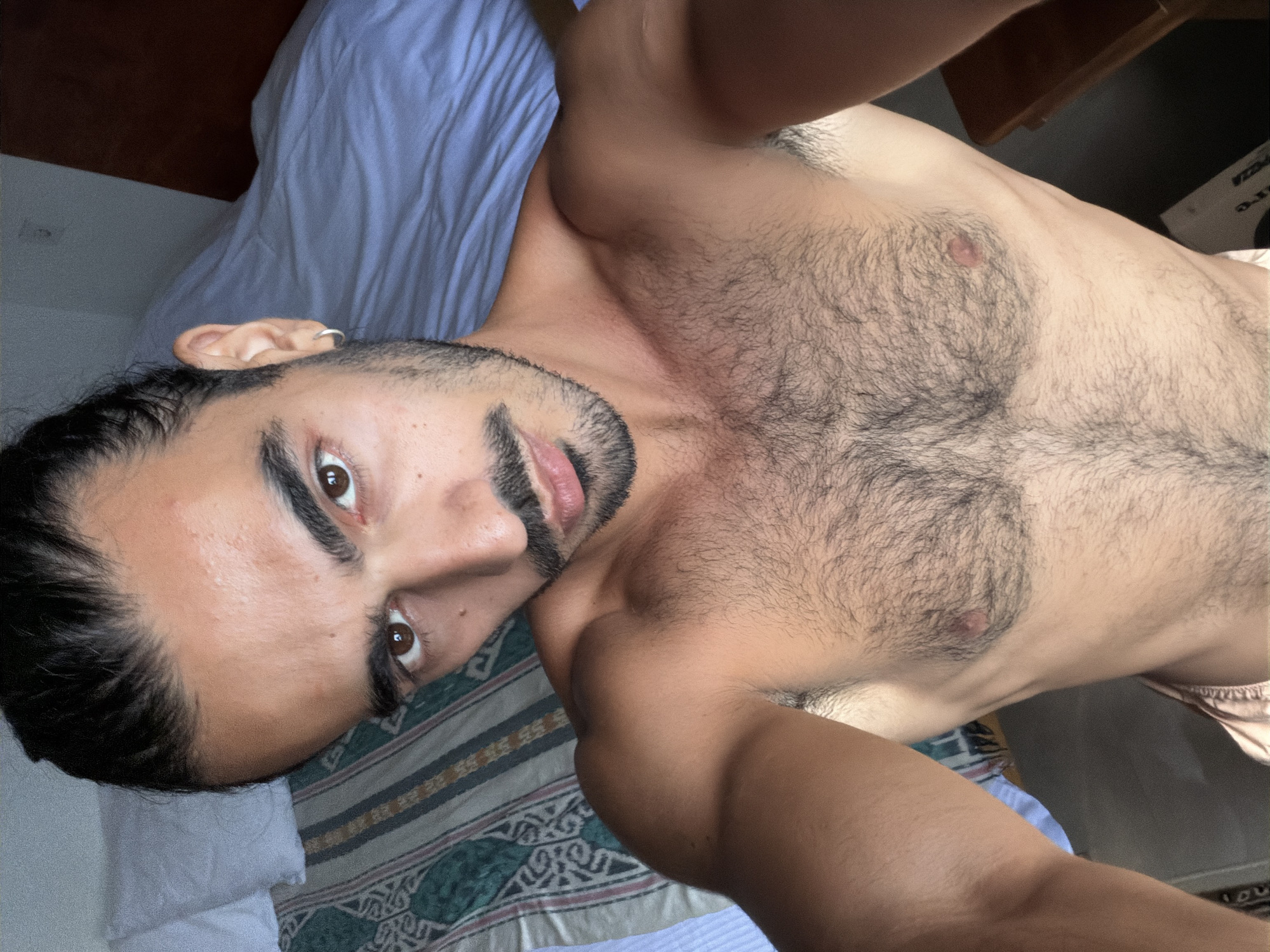 The media file displays a close-up, angled portrait of a man lying on a bed. He has dark hair, which appears to be styled in braids or small dreadlocks, a dark, well-maintained beard and mustache, and dark brown eyes that look directly into the camera. A small silver hoop earring adorns his left ear. He is shirtless, revealing a moderately hairy chest and bare arms.
The setting is indoors, presumably a bedroom, with the man positioned on a bed. The bedding consists of a light blue sheet or blanket and a patterned sheet underneath, featuring geometric designs in hues of green, blue, brown, and white. The lighting suggests daytime, with soft, natural illumination present. The man's expression is neutral and engaging. There is no visible text in the image. The location context provided is North Kuta, Indonesia.