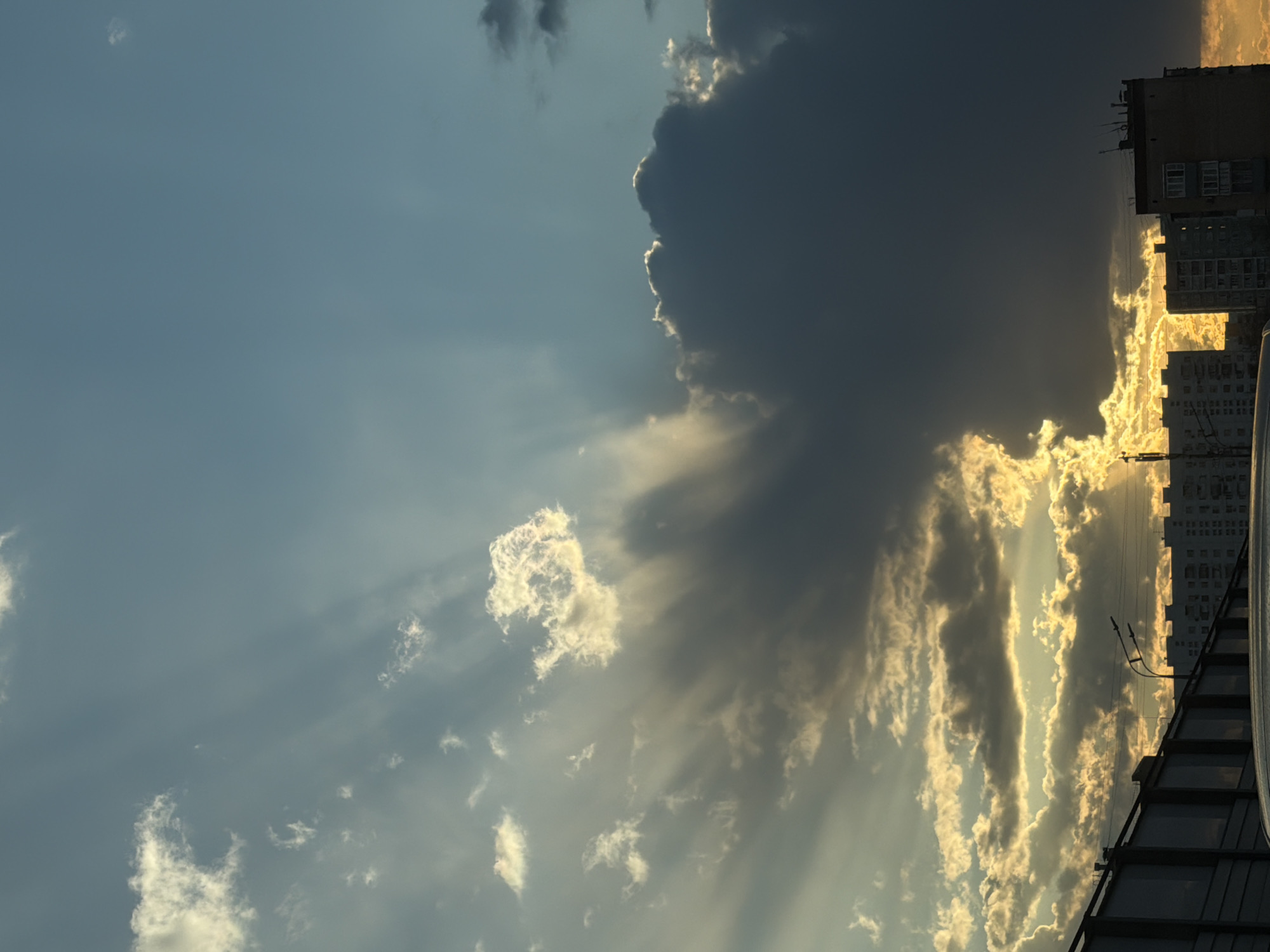 The image depicts a cityscape in Moscow, Russia, at dusk or dawn. The sky is mostly clear blue, with sunlight streaming through a large, dark cloud formation that dominates the right side of the frame. The sunlight is reflected and diffused, creating beams of light that illuminate the clouds from behind.
A tall building is visible on the right edge of the frame, partially obscured by the sunlit clouds. A rooftop structure is present in the bottom right corner. The scene suggests the intersection of natural beauty with urban development.