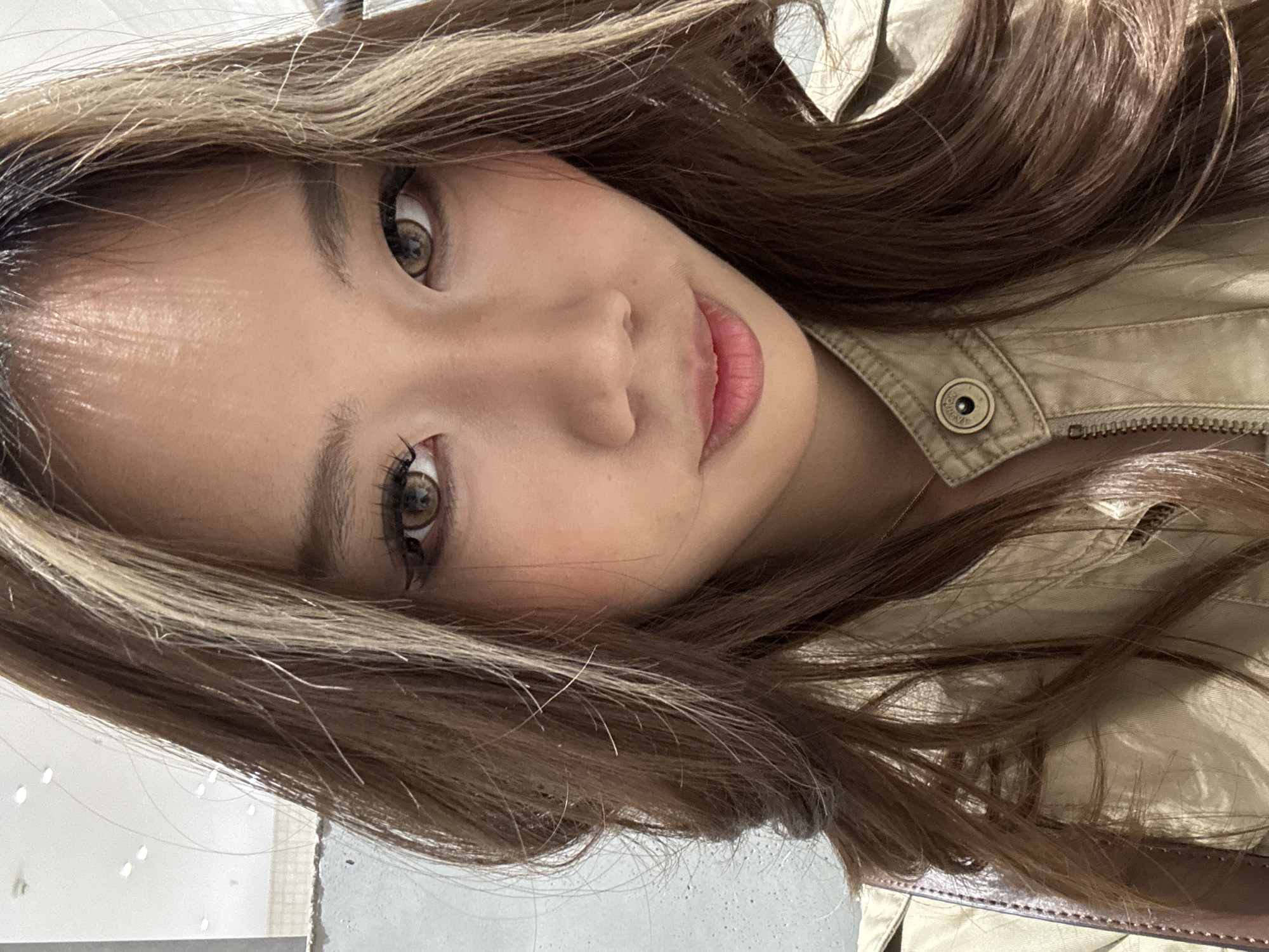 A close-up, slightly angled portrait shows a woman with light brown hair with blonde highlights. She has light skin, brown eyes, and light pink lips. She is wearing a khaki-colored jacket with a visible button and zipper. A glimpse of a white wall and a corner of an object, potentially a purse, are visible in the lower portion of the image. The location, Chenghua District, China, is not visually discernible from the image alone.