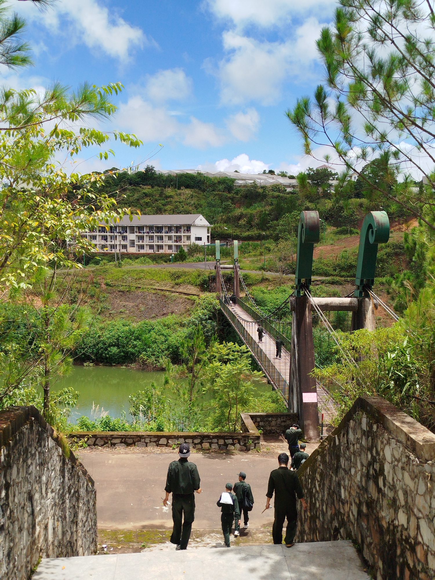 In Lạc Dương, Vietnam, a group of individuals in military uniforms are walking down stone stairs. The foreground is dominated by a rough-hewn stone wall on the right and a similar wall on the left, with concrete steps leading down. Several people in dark green uniforms are descending these stairs, with some carrying bags.
Further ahead, a suspension bridge spans a calm, green body of water. The bridge has a wooden walkway and is supported by two large, ornamental green pillars with spiral tops. A few individuals, also in military-style attire, are visible walking across the bridge. The surrounding environment is lush and green, with trees and dense vegetation on either side of the water.
In the background, a multi-story building with a light-colored facade and numerous balconies is situated on a gently sloping hillside. Above the building, several greenhouse structures are visible on the crest of the hill. The sky is a vibrant blue with scattered white clouds. The overall scene suggests a military or educational institution located in a natural, hilly landscape.