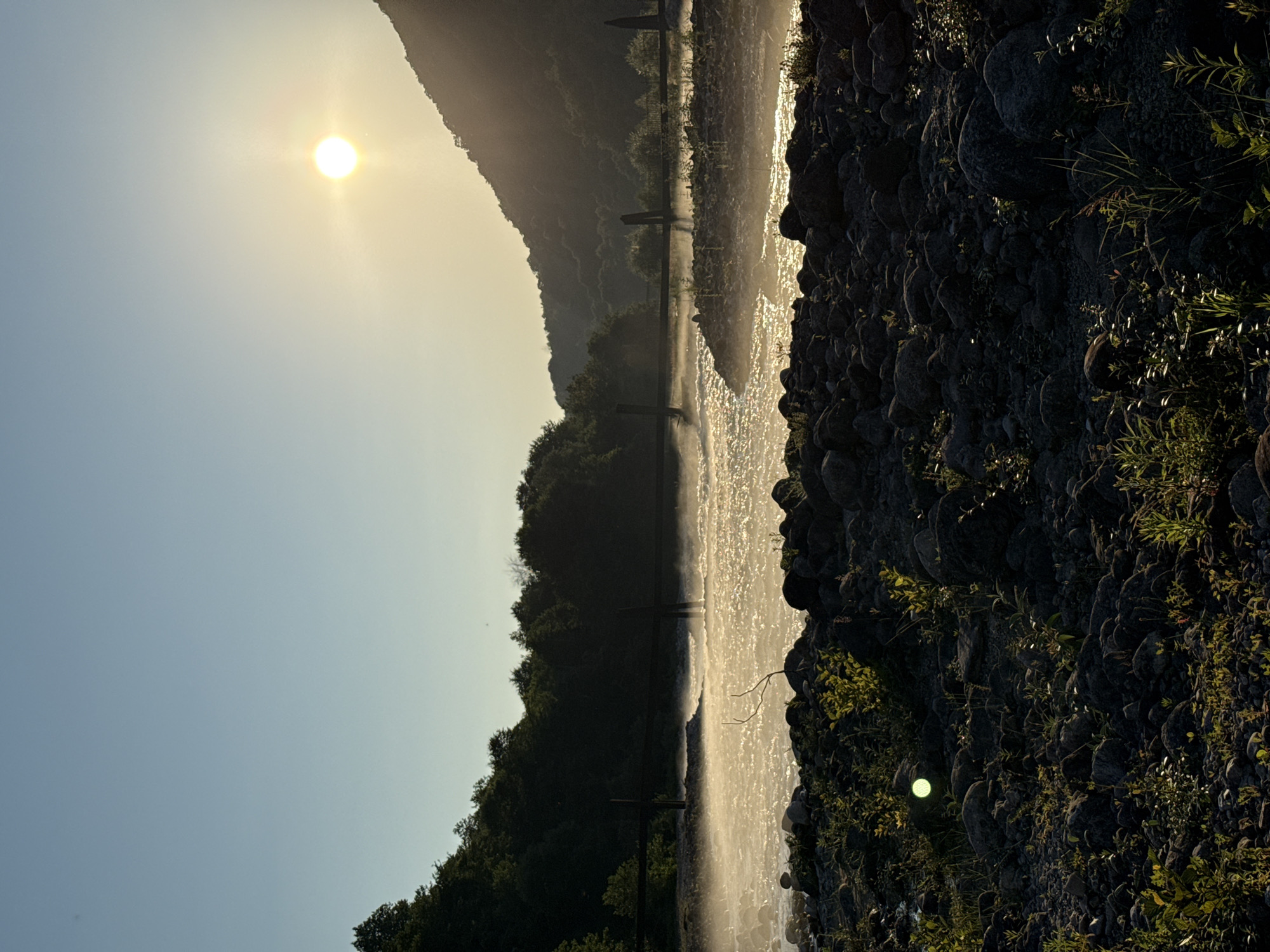 The image depicts a serene riverside scene at what appears to be sunset or sunrise, given the low angle of the sun casting a warm glow. The sun itself is a bright, diffused orb in the upper left portion of the frame, illuminating the sky with a pale yellow hue. The scene is oriented vertically, suggesting the photo was taken in landscape mode and then rotated.
The main subjects include a body of water, likely a river or stream, reflecting the sunlight and appearing shimmering and somewhat misty. Along the far bank of the water, a line of lush green trees and foliage forms a dense forest that climbs a gentle slope towards a silhouetted mountain range in the background. The presence of a few vertical posts or markers in the water suggests a shallow area, a weir, or a shallow crossing.
The foreground is dominated by a rocky shoreline, with large, dark stones and scattered green vegetation. The texture of the rocks is rough and uneven, with some smaller plants growing in crevices. The sunlight highlights the edges and surfaces of these rocks, creating a strong contrast with the shaded areas.
The overall mood is peaceful and tranquil, evoking a sense of natural beauty and quietude. The light is soft and diffused, contributing to the calm atmosphere. There is no visible text in the image. The location is identified as Abgara, Abkhazia. The weather appears clear, with the sun breaking through. No people or specific objects beyond the natural landscape elements and the suggestive posts in the water are identifiable.