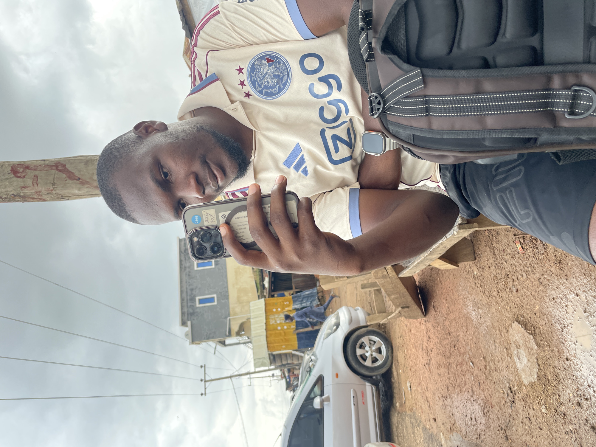 A man is taking a selfie with his phone outdoors. He is wearing a light-colored Ajax football jersey and black shorts. He has a backpack on his right shoulder. A smartwatch is visible on his left wrist. The background shows a cloudy sky, power lines, some buildings, and a white car. The ground appears to be dirt and is wet in some areas, suggesting it may have recently rained. The scene appears to be in a somewhat urban or semi-urban environment in Jos, Nigeria. The man's expression is neutral. The time of day is likely daytime due to the natural light. Text visible on the phone screen includes "256GB" and "100% PASSED". The Ajax logo and the "A" logo of Adidas are visible on his jersey.