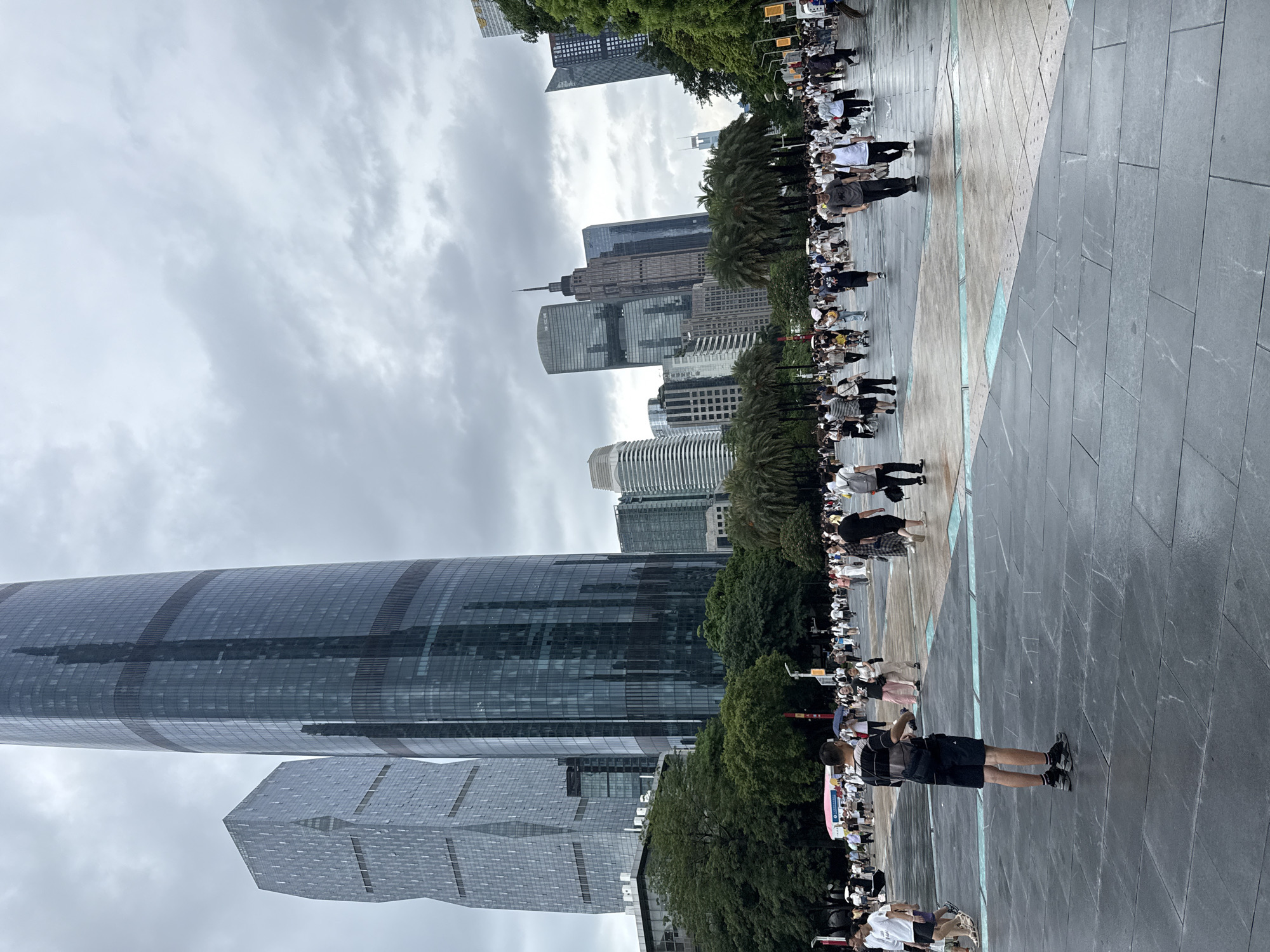 A street scene in Tianhe District, China depicts a cloudy day with a large crowd of pedestrians in an outdoor plaza. In the background, several modern skyscrapers dominate the skyline. The foreground shows a man standing with his back to the camera, wearing a striped t-shirt, black shorts, and sneakers. He is looking towards the crowd. The plaza is paved with dark gray tiles and has some colored accents. People of all ages are walking around, some with umbrellas, suggesting a possibility of rain or recent rain. The overall atmosphere is bustling and urban.