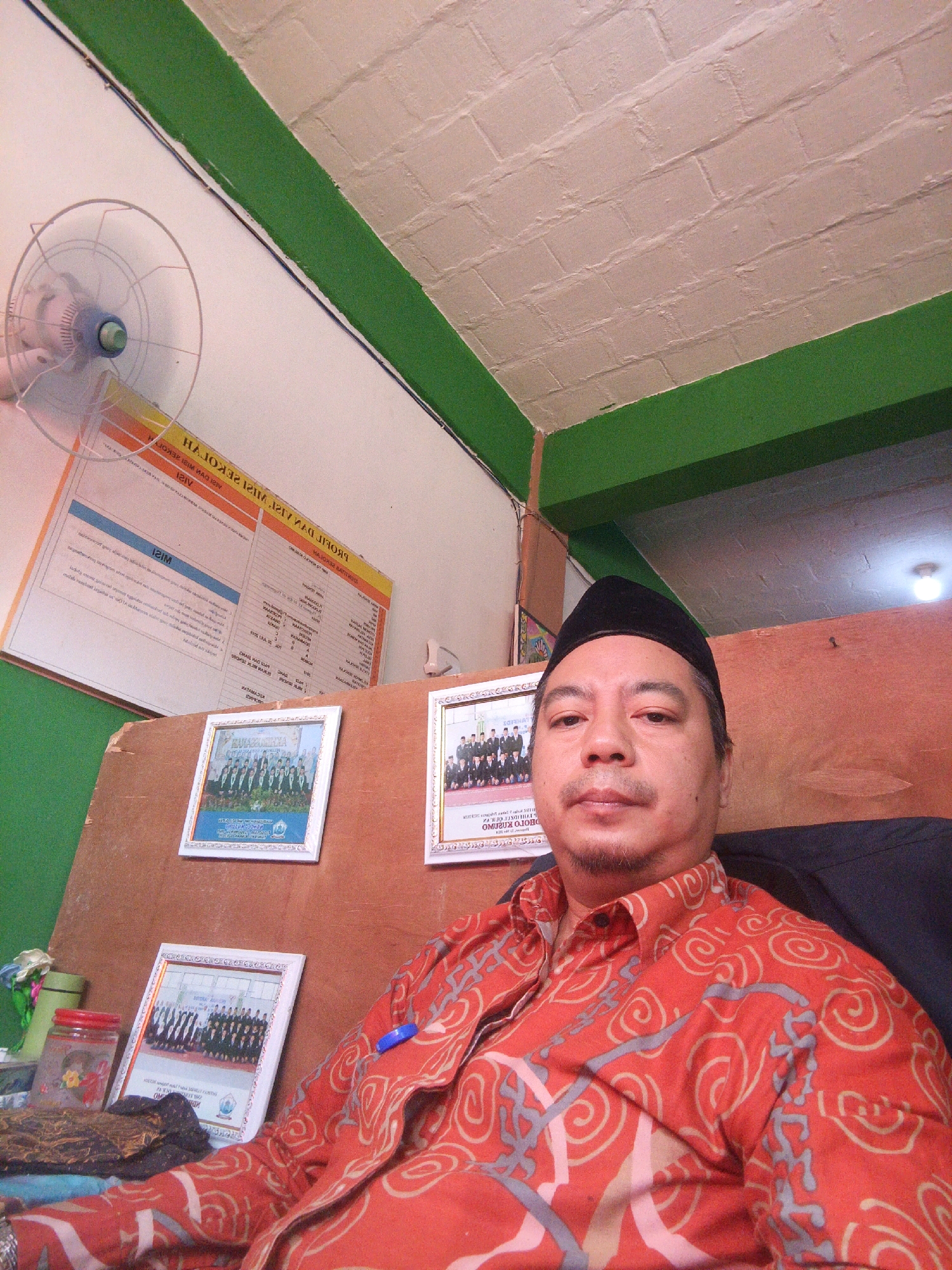The image captures a man, likely Indonesian, posing for a selfie. He is wearing a black peci cap and a red batik shirt with a swirling pattern. The man is seated, with his upper body filling the foreground. Behind him, a wall is visible, adorned with framed photographs, suggesting a personal or professional space. One framed photograph displays a group of people in uniform, possibly students or staff. Another photograph shows a similar group, hinting at an institutional setting. A whiteboard or poster is also partially visible on the wall, featuring text in what appears to be Indonesian. A ceiling fan is mounted on the wall in the upper left corner. The overall environment appears to be an indoor space, possibly an office or classroom, with green painted accents on the wall and ceiling. The lighting is moderately bright, illuminating the scene. The background hints at a simple, functional setting, typical of a school or community building. There are no specific indicators to pinpoint the location within Tlogowungu, Indonesia, beyond the context of Indonesian language and cultural attire.