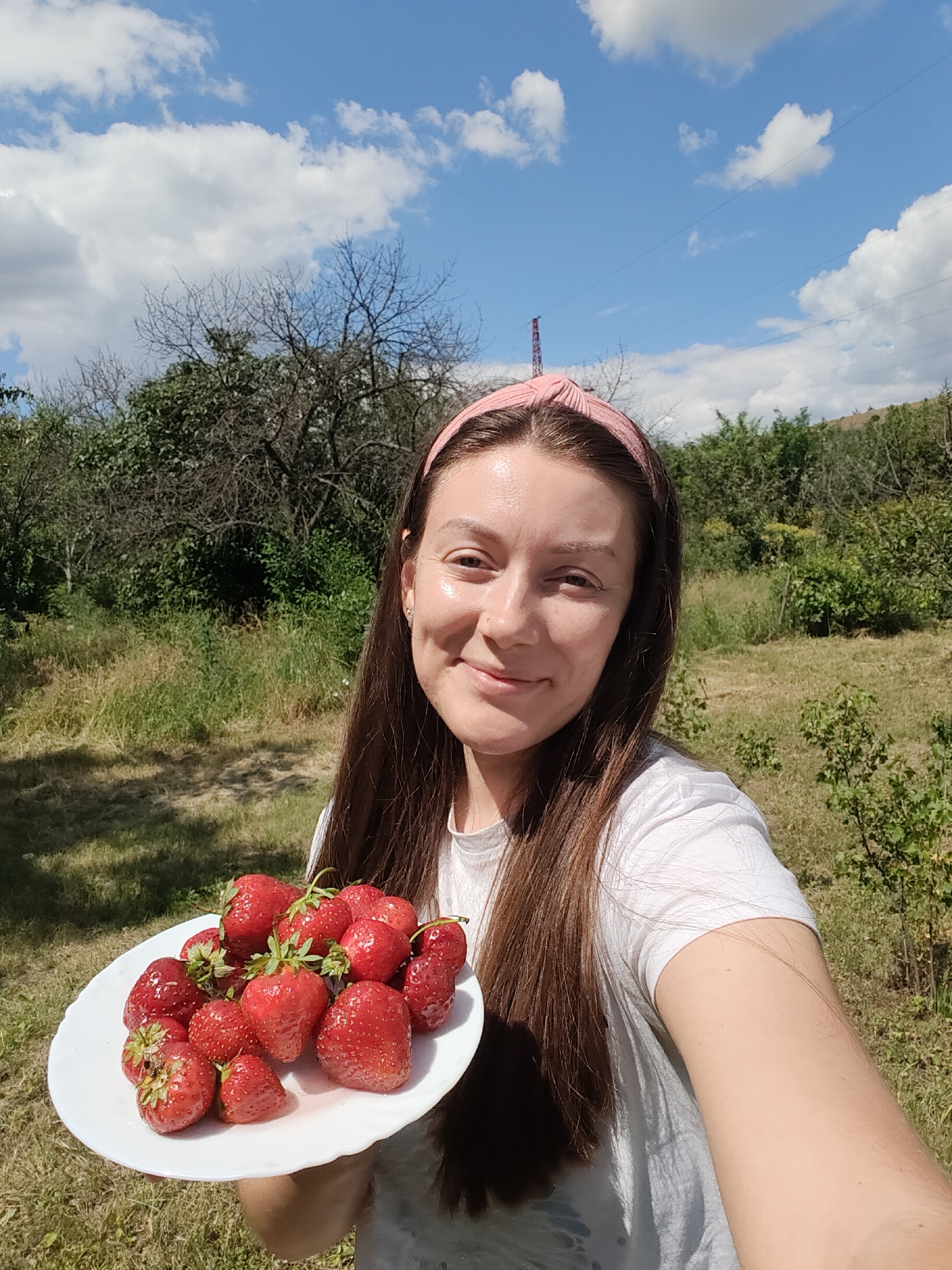 A young woman with long brown hair and a pink headband is taking a selfie outdoors. She is smiling and holding a white plate filled with fresh strawberries. The background shows a lush green landscape with trees, grass, and a bright blue sky with white clouds. There is a power line visible in the distance, and a metal tower. The woman is wearing a white t-shirt. The image appears to have been taken on a sunny day. The location is the city of Kryvyi Rih, Ukraine.