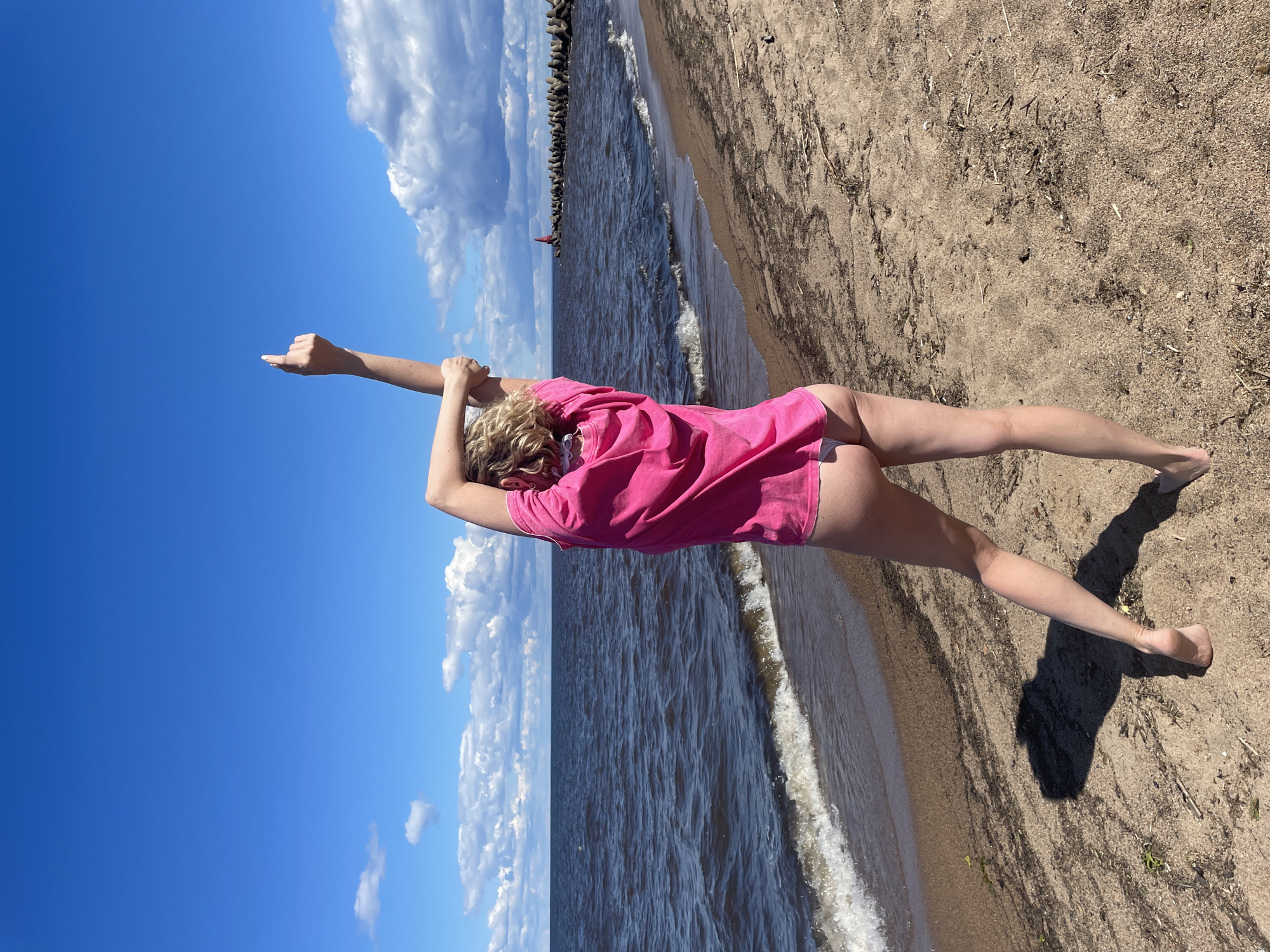 A woman with blonde curly hair stands on a sandy beach, facing away from the camera. She is wearing a bright pink t-shirt and white bikini bottoms. Her left arm is raised above her head, with her hand pointing upwards. Her right arm is bent, with her hand resting on her head. The woman is positioned in the lower right quadrant of the image, with her shadow cast on the sand to her left.
The beach is composed of light brown sand with scattered pieces of seaweed and debris. Gentle waves from a dark blue body of water lap at the shore. In the background, a breakwater made of concrete tetrapods stretches into the water, with a red beacon visible at its end. The sky is a vibrant blue, dotted with fluffy white clouds. The overall scene suggests a sunny day at the coast. This location is likely the shoreline of Lake Beloye in Belozersk, Russia, given the geographical context.