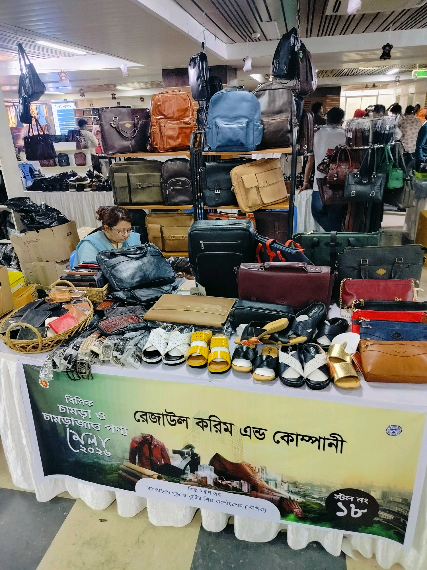 The image depicts a stall at the "Dhaka Leather Goods and Footwear Fair 2022" (Dhaka Charmra O Charmrajato Ponnyo Mela 2022) held in Dhaka, Bangladesh. The stall is covered by a white cloth draped table, upon which a variety of leather goods and footwear are displayed. Items include handbags, backpacks, wallets, and various styles of sandals and shoes, in colors such as black, brown, tan, red, blue, green, and white. Some items are presented in wicker baskets or on shelves behind the table. A woman wearing a blue hijab and glasses is seated behind the table, appearing to attend to the display. In the background, other stalls and shoppers are visible, along with signage and lighting fixtures suggesting an indoor exhibition hall. The banner at the front of the stall features Bengali text and graphics of a shoe and city skyline, indicating the name of the participating company as "Regalium Karim and Company" and its involvement with the Ministry of Industries. The overall scene is one of a commercial exhibition focused on leather products.