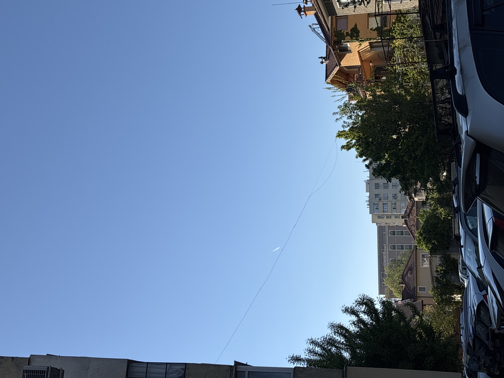 The image shows a street scene in an urban environment. Buildings of varying heights and architectural styles line the street, some with trees in front. A row of parked cars is visible on the right side. Overhead, a thin wire or cable stretches across the blue sky, with what appears to be an airplane or contrail visible near the wire's path.