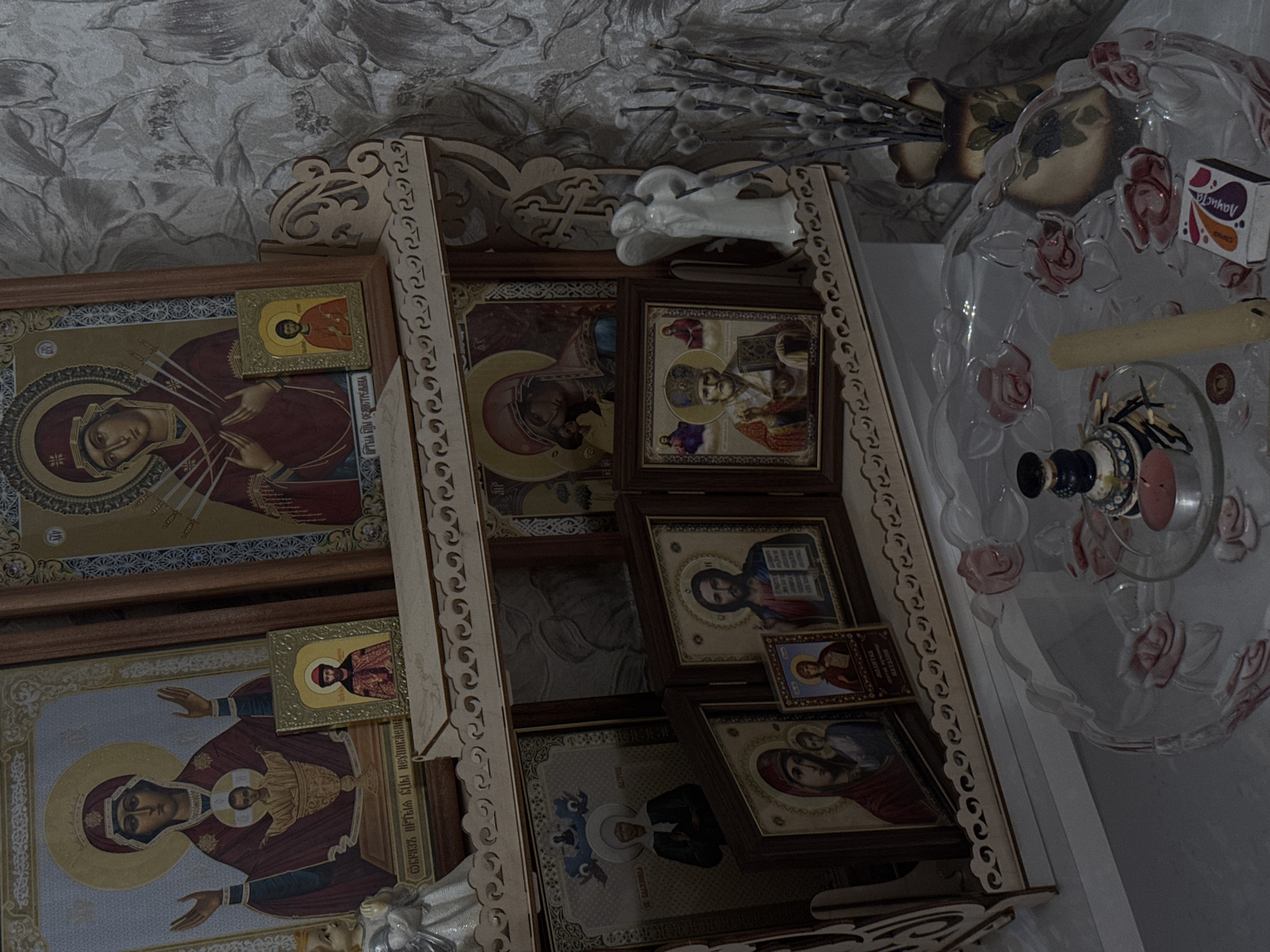 The image depicts a collection of Orthodox Christian icons displayed on a wooden shelf with decorative cutouts. The icons vary in size and depict various religious figures. On the top shelf, there are multiple icons, including one featuring Mary and Jesus. An angel figurine and a vase with willow branches sit atop the shelf. To the right of the shelf, there's a glass dish with floral decorations, a box of matches, a candle, and other small items. The background features a wallpaper with a floral pattern. The location appears to be a domestic interior, possibly in a home in an unknown city.