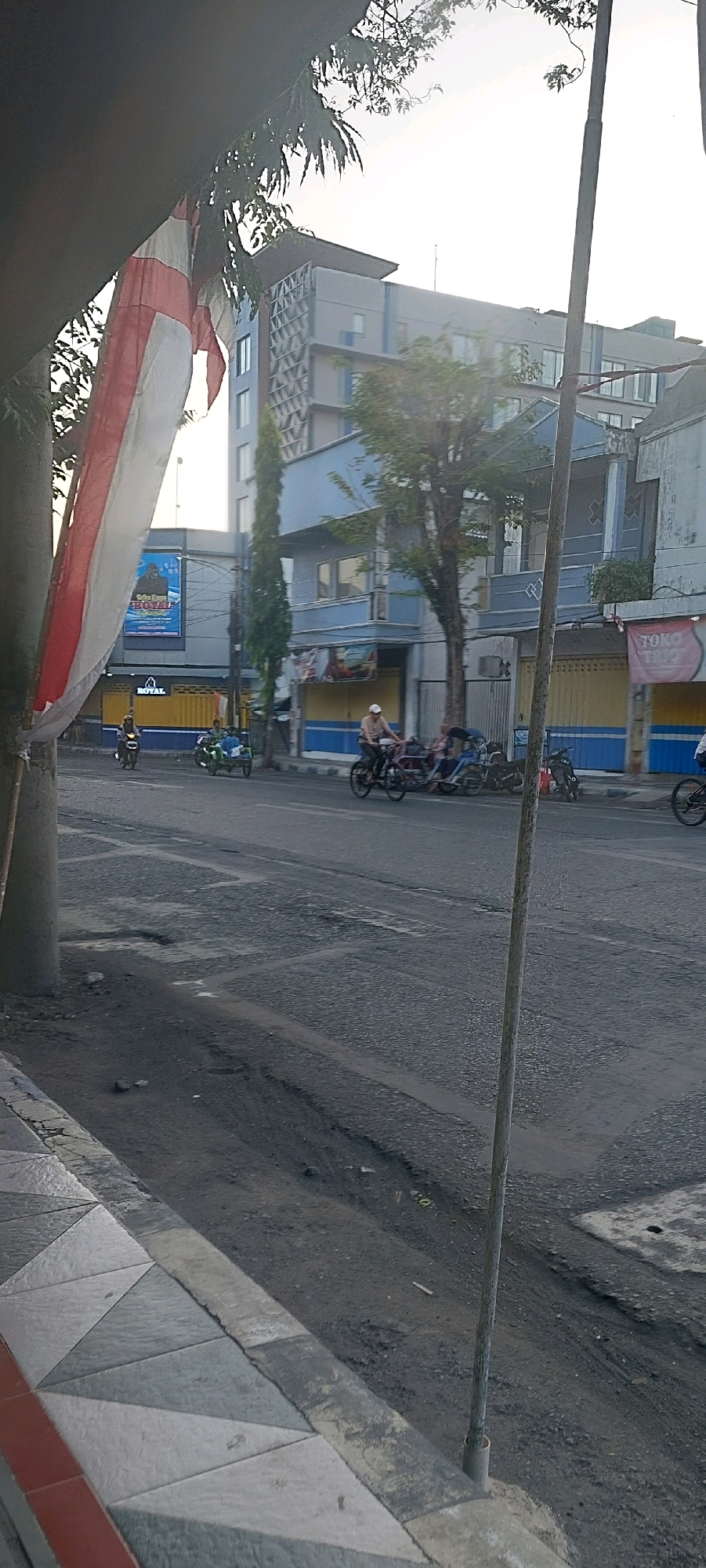 In Lumajang, Indonesia, a street scene unfolds. A white and red Indonesian flag hangs from a pole on the left, partially obscuring a modern, multi-story building with a patterned facade. The building appears to be a hotel or commercial establishment, with a blue and yellow striped awning visible on a lower level. Across the street, a two-story building with blue accents and yellow roller doors is situated.
Several individuals are present on the road. A person rides a motorcycle from left to right. Further down the street, a green tricycle-like vehicle carries two passengers, and behind it, a person on a bicycle pulls a similar passenger cart. Another motorcycle is parked to the right of the blue-accented building. The road surface is asphalt and shows signs of wear with visible potholes and repairs. A thin, weathered pole stands vertically in the foreground, and to the left, a patterned sidewalk made of concrete tiles is visible. The sky is bright, suggesting daylight, possibly late afternoon due to the angle of the light.