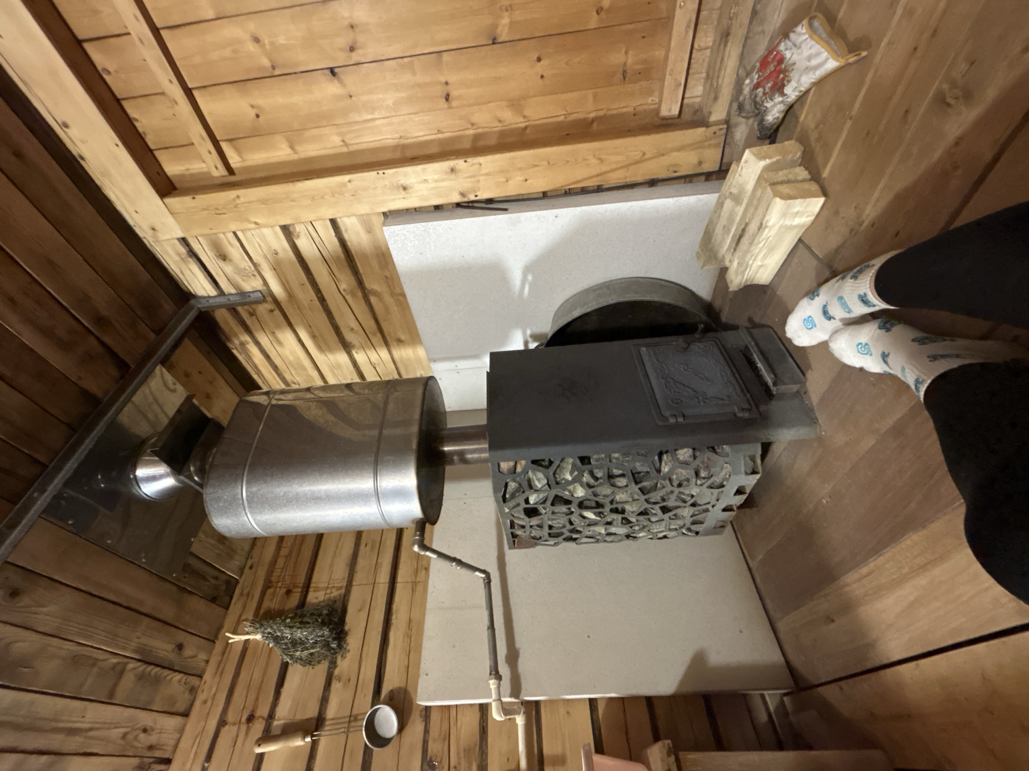 An interior shot shows a sauna stove with a cylindrical metal water tank connected by pipes, surrounded by wood paneling. A metal scoop, birch branches, and a glove sit near the stove. Wood logs are stacked beside the stove. Two people in socks and black pants stand near the stove. The city of Bashur, Russia is not discernible from the image.