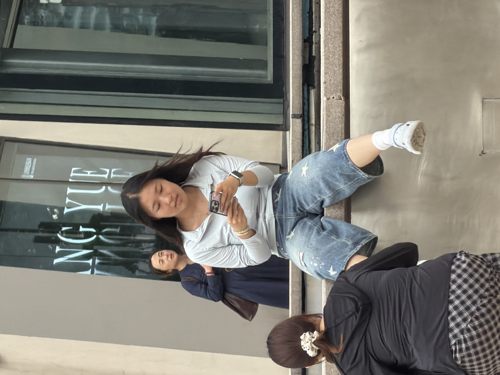 In Yuzhong District, China, a young East Asian woman is seated on a ledge, engrossed in her smartphone. She is wearing a light blue zip-up hoodie and distressed denim shorts adorned with white stars. Her right leg is crossed over her left, and she is wearing white socks and light blue clogs. To her left, another East Asian woman in a dark blue dress stands facing away from the camera, holding a brown bag over her shoulder. In the foreground, partially visible, is the back of a third East Asian woman wearing a black hooded jacket and a plaid skirt. The background features a building with large glass windows reflecting the words "NG YUE". The scene appears to be a public outdoor space, possibly a plaza or walkway, with architectural elements suggesting an urban setting.