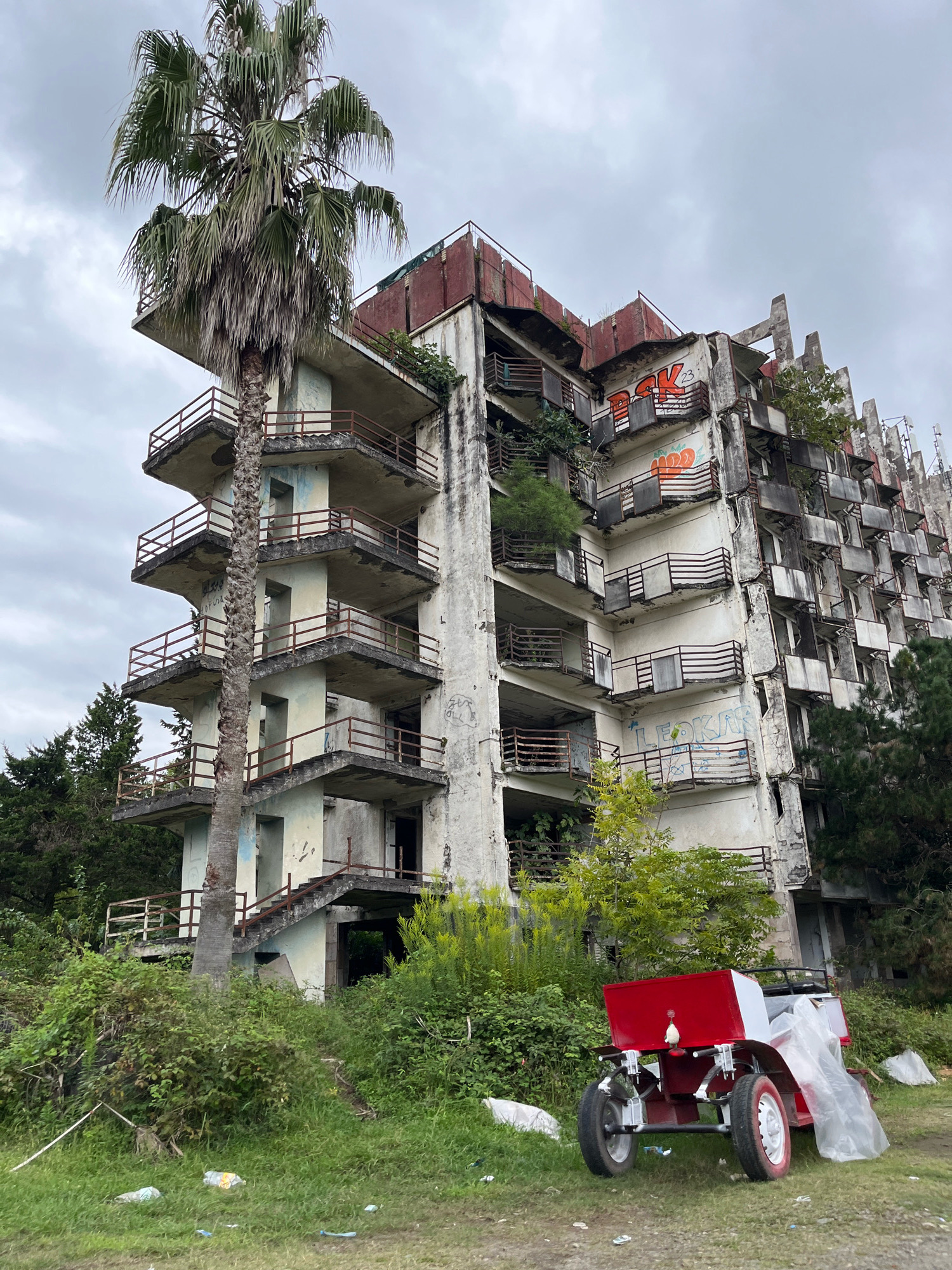 An abandoned, multi-story building with a brutalist architectural style stands on the outskirts of Ureki, Georgia. The building is in a state of severe disrepair, with crumbling concrete, rusting balconies, and overgrown vegetation. Graffiti covers some of the exterior walls. In the foreground, a small, red and white cart with two wheels is parked on a grassy area littered with trash. A tall palm tree stands to the left of the building, its fronds partially obscuring the structure. The sky is overcast, casting a somber mood over the scene. The overall impression is one of decay and neglect.