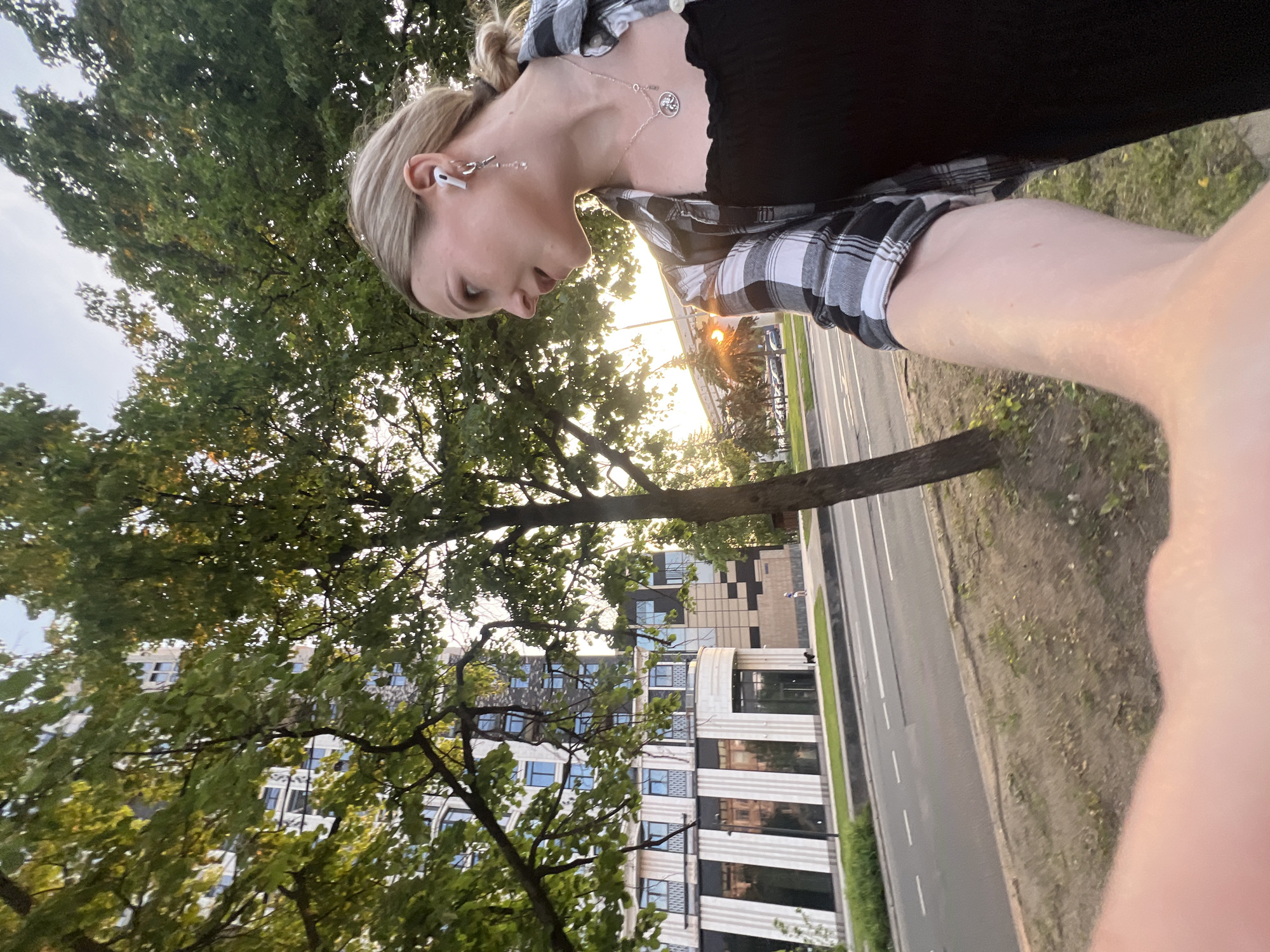 A young Caucasian woman with blonde hair pulled back in a ponytail is walking outdoors in the city of Khimki, Russia. She is wearing a black off-the-shoulder top and a black and white plaid shirt, and has a necklace with a circular pendant. She is also wearing white wireless earbuds and a dangling earring. The woman is looking to her right, and her arm is extended forward, as if she is taking a selfie.
In the background, a modern apartment building with a facade of white stone and dark brick is visible. The building has numerous windows and balconies. A street with cars is in front of the building, and trees line the sidewalk. The sun is setting, casting a warm glow on the scene. There is a small black dog on the grass in front of the building.