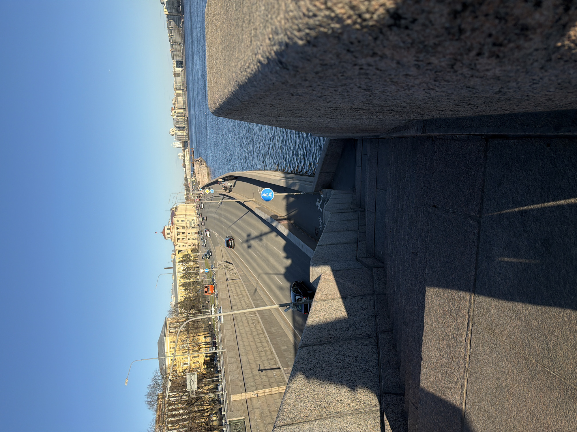 This is an elevated perspective of a section of Liteyny Bridge in Saint Petersburg, Russia. The photograph is taken from the bridge's edge, showing a view of the roadway below and the Neva River in the distance. The bridge's stone construction is prominent in the foreground, with its textured surface and shadows cast by sunlight. The roadway features light automobile traffic and pedestrian walkways with visible road markings and signage, including a designated bicycle path. Buildings line the far side of the river, visible against a clear blue sky. The lighting suggests it is daytime with clear weather. The composition is slightly tilted, altering the orientation of the scene.