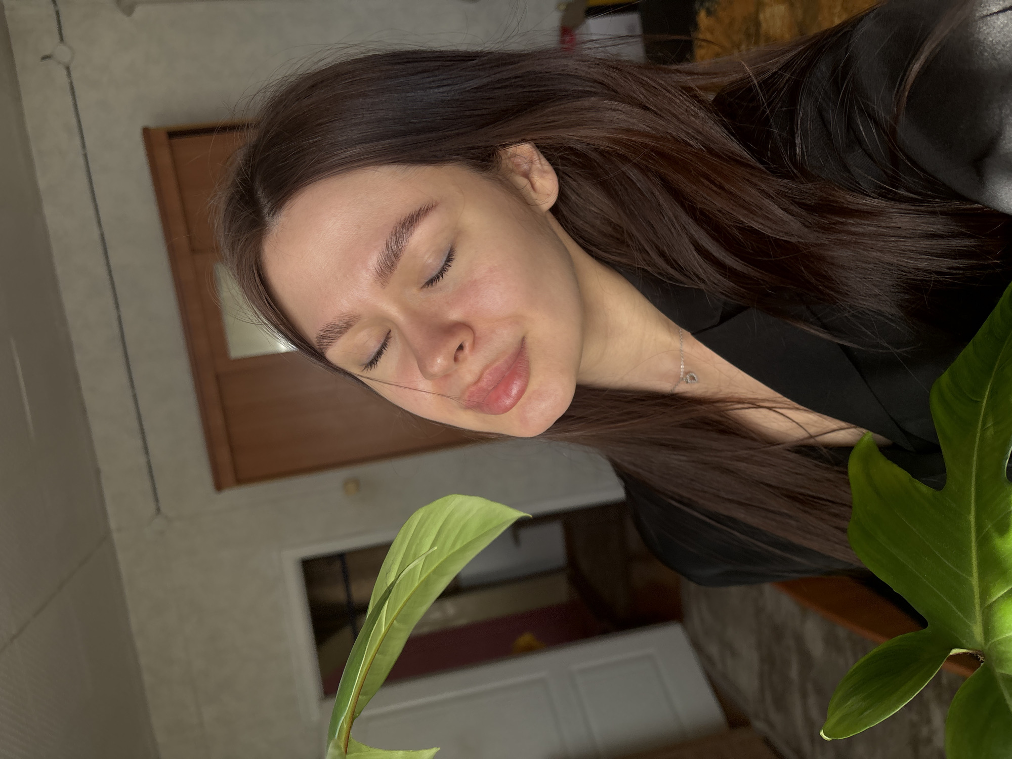 A woman with long, dark hair is the central subject. Her eyes are closed, and she is wearing a black garment, possibly a jacket or robe. She is wearing a delicate necklace. In the foreground, there is a large, green plant with prominent leaves. The setting appears to be indoors, evidenced by a wooden door and neutral-colored wall in the background. The lighting is soft and diffused. The image does not provide enough information to ascertain if the photo was taken in Dubrova, Russia.