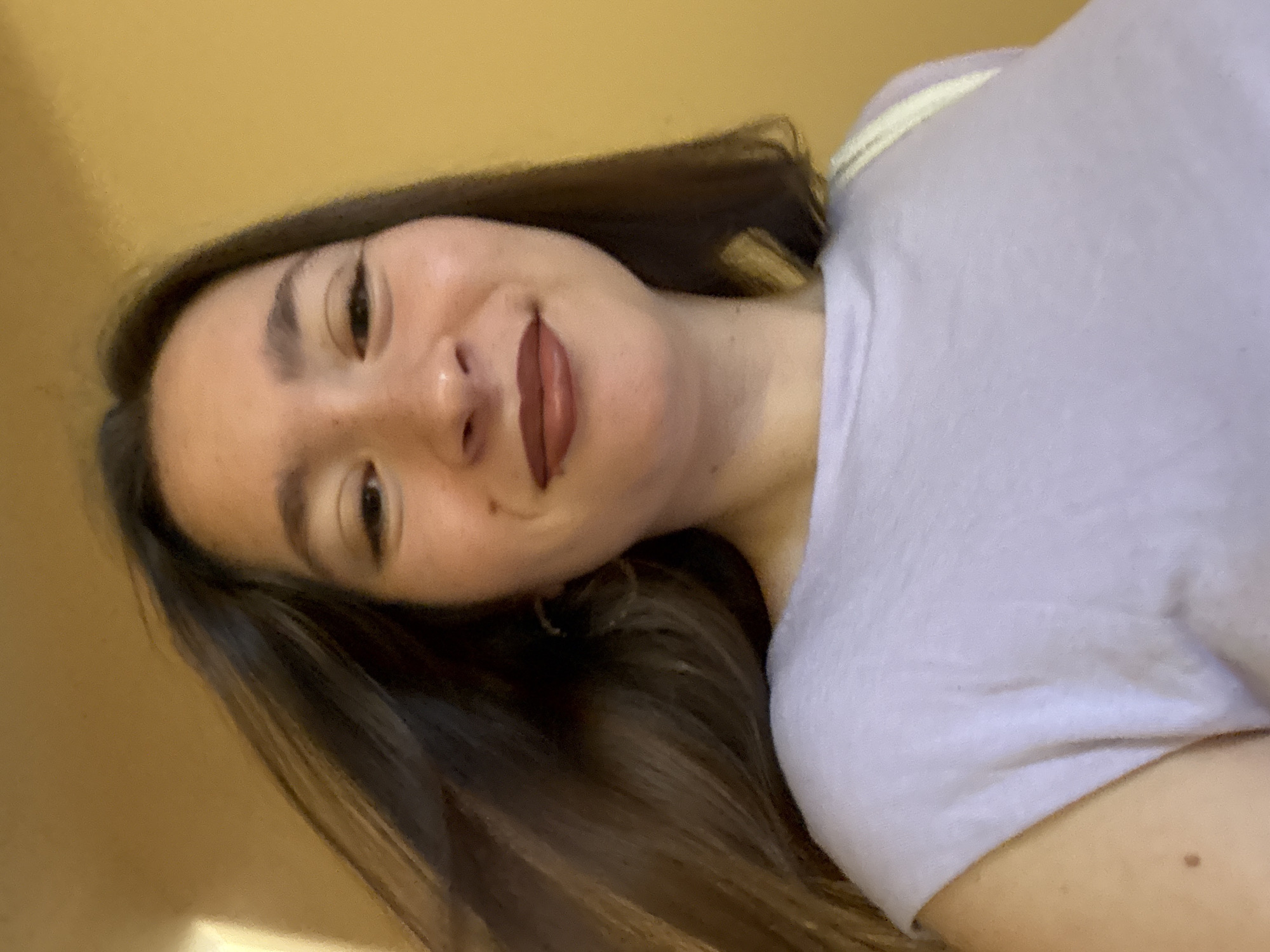 A young woman with long, dark brown hair is looking into the camera, her head tilted slightly. She is wearing a light purple t-shirt. Her lips are painted with a dark reddish-brown lipstick. There's a light colored strap visible on her right shoulder, possibly from a bag. The background is a solid, warm yellow wall, with a hint of a lighter wall or doorway on the far left. The lighting appears to be natural, coming from the left side of the frame. The overall impression is of a casual, everyday moment captured in Sofia, Bulgaria, perhaps in an interior residential setting or a building hallway.