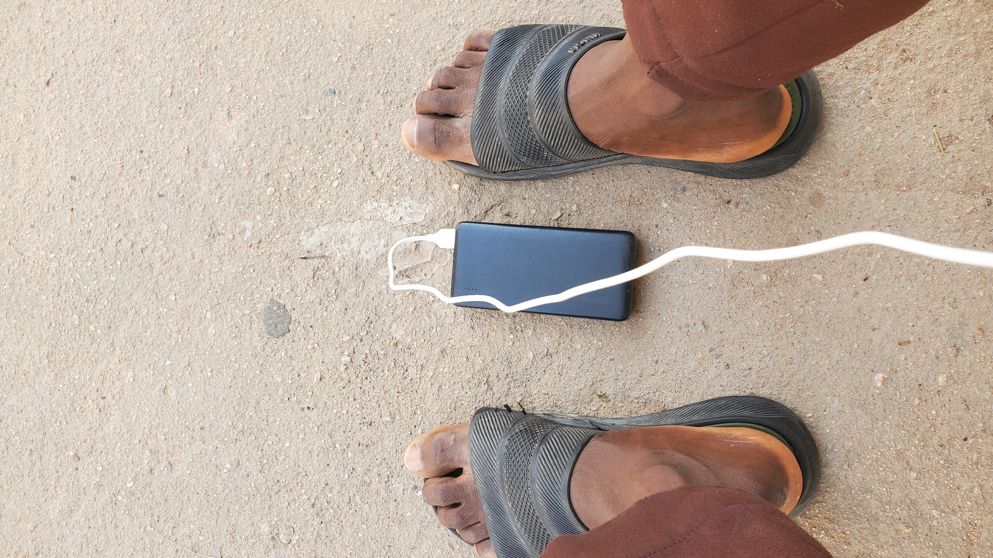 An overhead shot shows two bare feet wearing black Adidas slides on a sandy ground, one on each side of a black power bank charging a device, with a white charging cable connecting them. The scene is set in Jalingo, Nigeria.