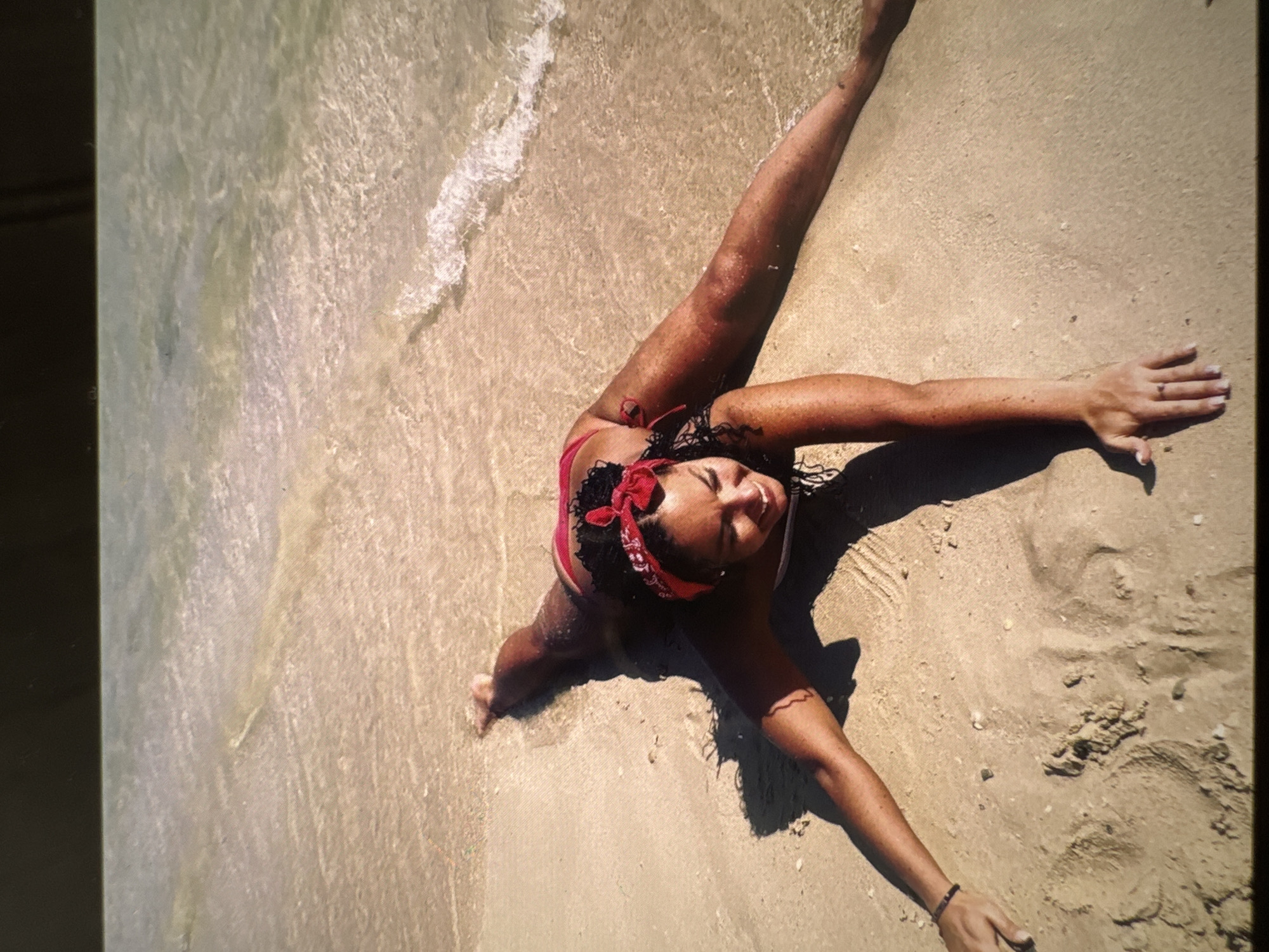 The image shows a woman lying on a sandy beach, with the water lapping at her feet. She is wearing a red bandana and a bikini. The woman is smiling and appears to be enjoying herself. The beach is clean and there are a few small shells scattered on the sand. The water is clear and shallow. The lighting suggests it is daytime. The image does not provide any information that would allow it to be definitively located in Sochi, Russia.