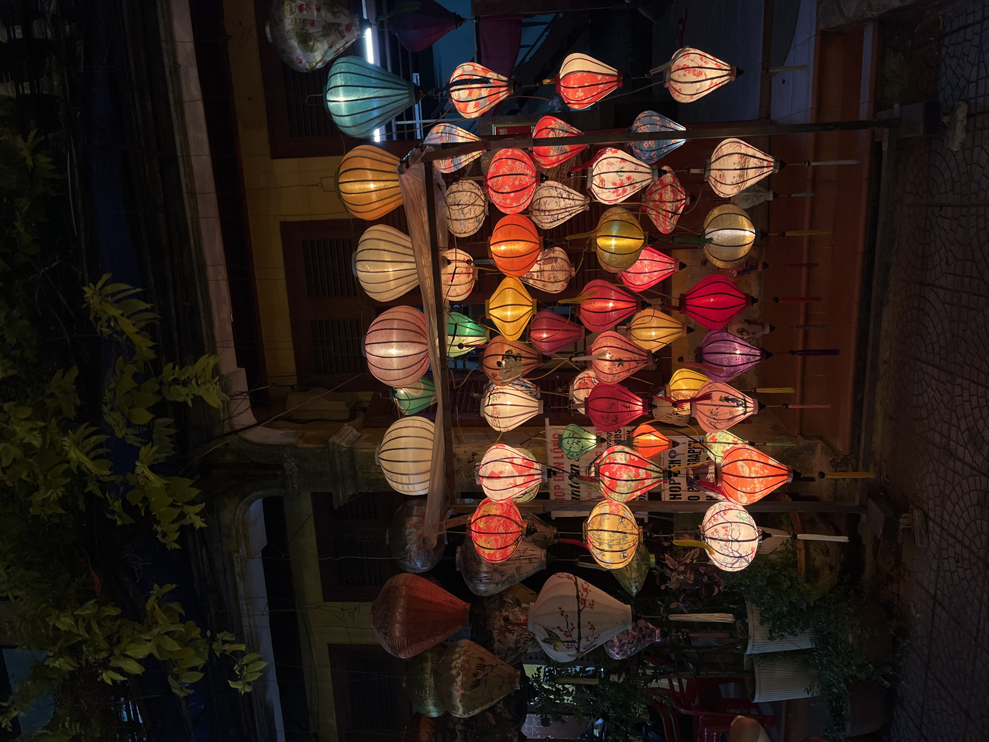 The image shows an outdoor scene in Hội An Ward, Vietnam, at night. Several rows of colorful, illuminated silk lanterns hang from a horizontal pole structure that is affixed to the sides of buildings. The lanterns are in a variety of shapes including round and oval with striped, floral, and solid designs, ranging in colors such as blue, red, pink, orange, yellow, green, purple and white. The buildings on either side of the lanterns feature closed shutters and other architectural details visible in the dim light. A tree branch with foliage is visible on the left side of the frame. Potted plants and red chairs are seen at the bottom right of the image. A stone paved road is seen on the right and dark patches of ground are seen on the left.