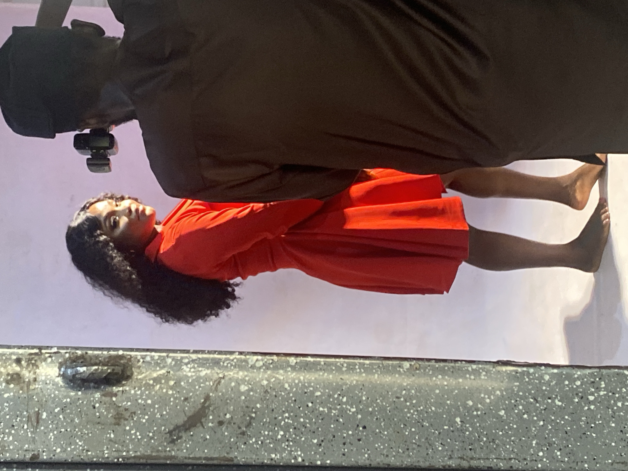 The image shows a woman in a red dress with dark curly hair, posing in an indoor setting with a light pink or off-white wall as a backdrop. A person in dark clothing stands partially in the upper section of the frame, facing downwards, with what appears to be a camera attached to their head or chest. The woman is barefoot. The bottom part of the frame shows a section of a gray surface speckled with white dots, potentially a table or ledge. There is insufficient information to determine the precise location in Jos, Nigeria. The scene suggests a photoshoot or video recording session.