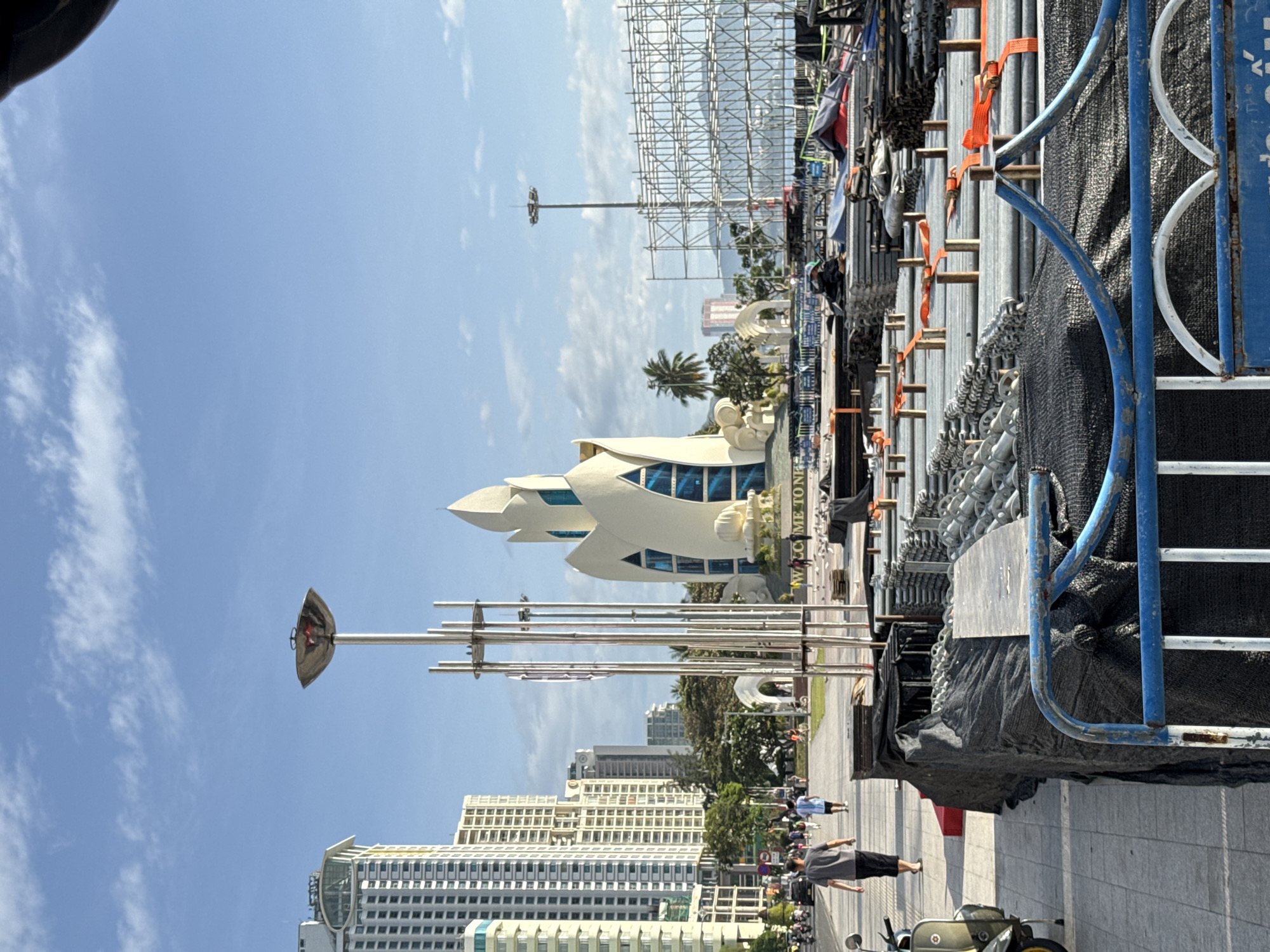 This image captures a street-level view of a scenic waterfront area in Nha Trang, Vietnam. The scene is dominated by a large, distinctive white building with a unique, petal-like architectural design, resembling a lotus flower. In the foreground, the pavement is lined with stacks of construction materials, including metal scaffolding and pipes, covered in black netting and some orange straps, indicating ongoing or recently completed construction or event setup.
To the left of the lotus-shaped building, several tall, modern high-rise buildings rise into the clear blue sky, typical of a coastal city. A pedestrian walkway is visible, with several individuals walking along it. A man in a grey t-shirt and black shorts walks away from the camera, and other people are seen further down the walkway. A vintage-style scooter is parked on the left side of the walkway.
In the middle ground, a tall, modern street lamp with a wide, bowl-shaped fixture stands prominently. Behind the construction materials and the walkway, a body of water, likely the sea, is visible in the distance, with a clear horizon under a partly cloudy sky. A large metal scaffolding structure, possibly for an event or display, is erected on the right side of the frame, further emphasizing the presence of construction or preparations. The overall impression is a blend of urban development, public space, and construction activity in a popular tourist destination.