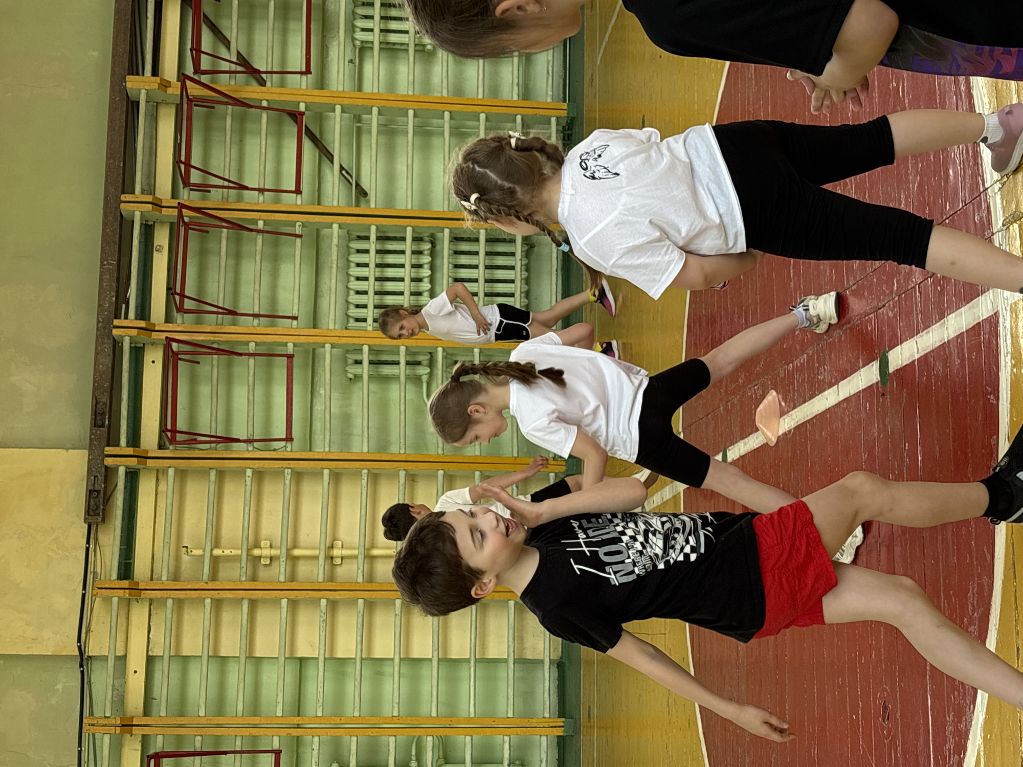 The image depicts a group of children engaged in some kind of physical activity inside a gymnasium, possibly located in Izhevsk, Russia. The children are wearing a mix of casual clothing including t-shirts, shorts, and leggings. They are positioned on a gymnasium floor with red and white lines. In the background, there are wall bars, a common feature in school gyms. The walls of the gym are painted in light shades of green and yellow. The activities the children are involved in appear to be part of a school gym class or an extracurricular activity.
