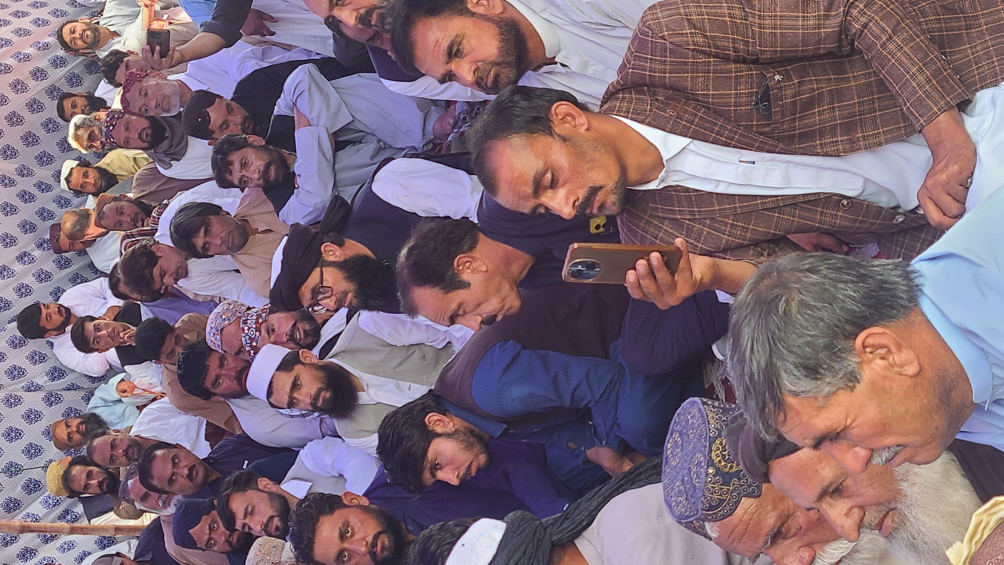A large group of men are seated on a patterned surface, possibly a rug or blanket, in Panjpai, Pakistan. Many are wearing traditional clothing. One man is holding and looking at a smartphone. Another man in a plaid jacket is looking down. Some men are wearing hats or head coverings. One man is kissing the cheek of another man with a white beard. The overall scene suggests a gathering or meeting.