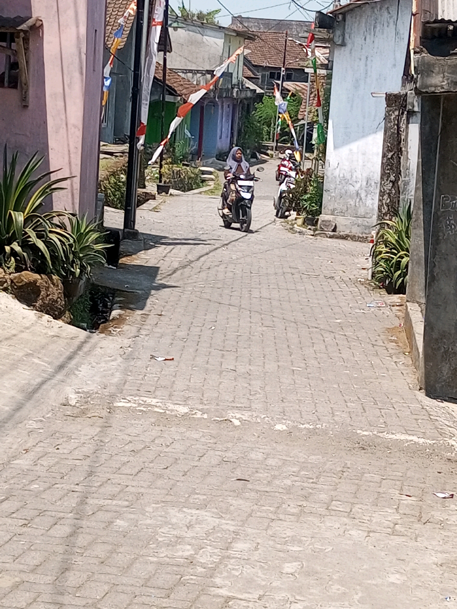 In the city of Andongsili, Indonesia, a paved street in a residential area is depicted. The street is lined with buildings, some with weathered facades. Small flags, possibly national or festive decorations, are visible on poles and attached to buildings. Two individuals are riding motorcycles on the street, traveling in the same direction. The foreground shows a portion of the paved road with some debris, while the background features more of the street and buildings under a bright sky.