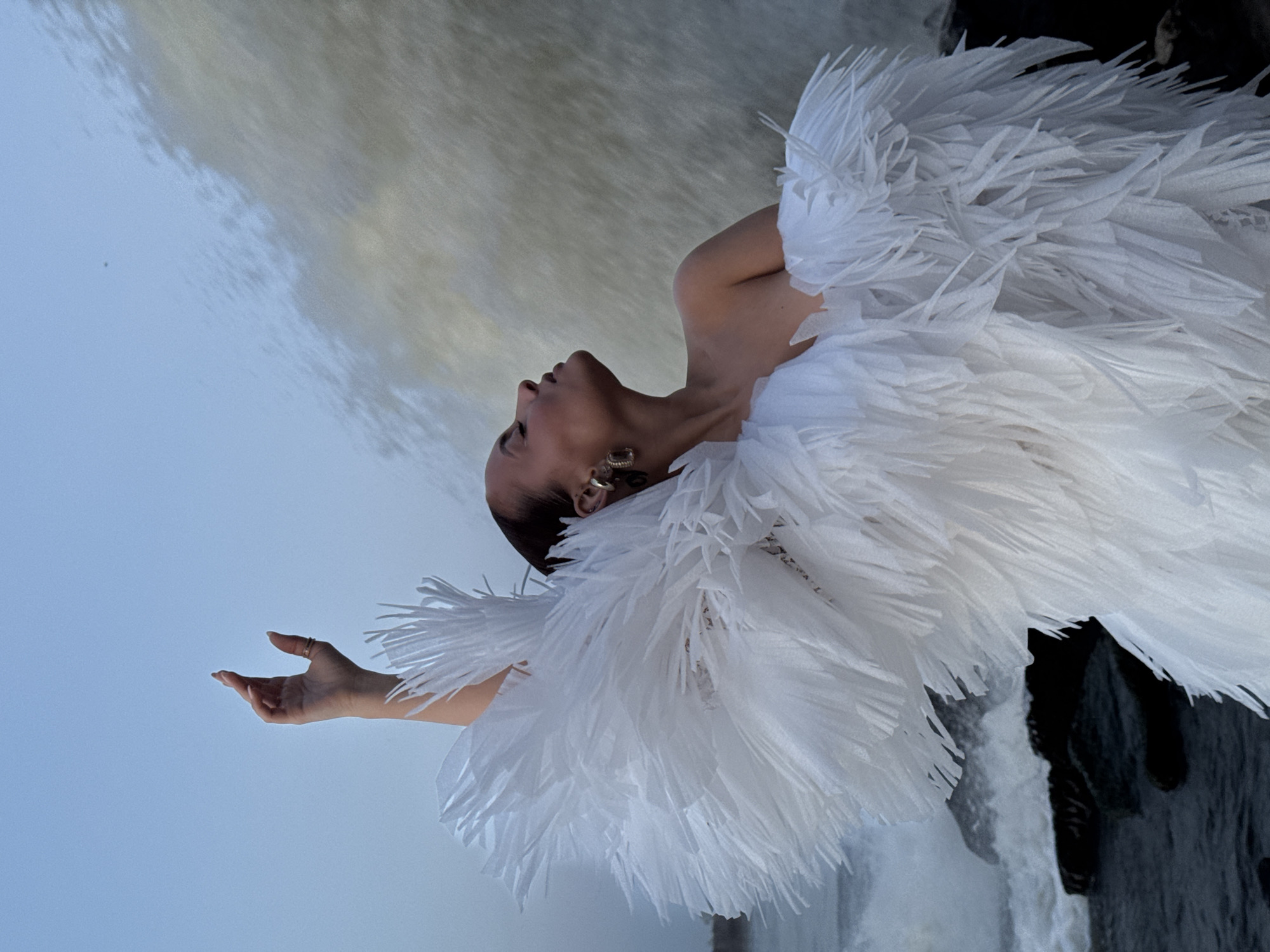 A woman with a shaved head is wearing a white feathered costume, with a waterfall in the background. The woman is facing upwards, her right arm extended, her left arm is visible with her earrings. The location is likely in Chaikovsky, Russia given the context. The environment consists of the waterfall, water spray and dark rocks.