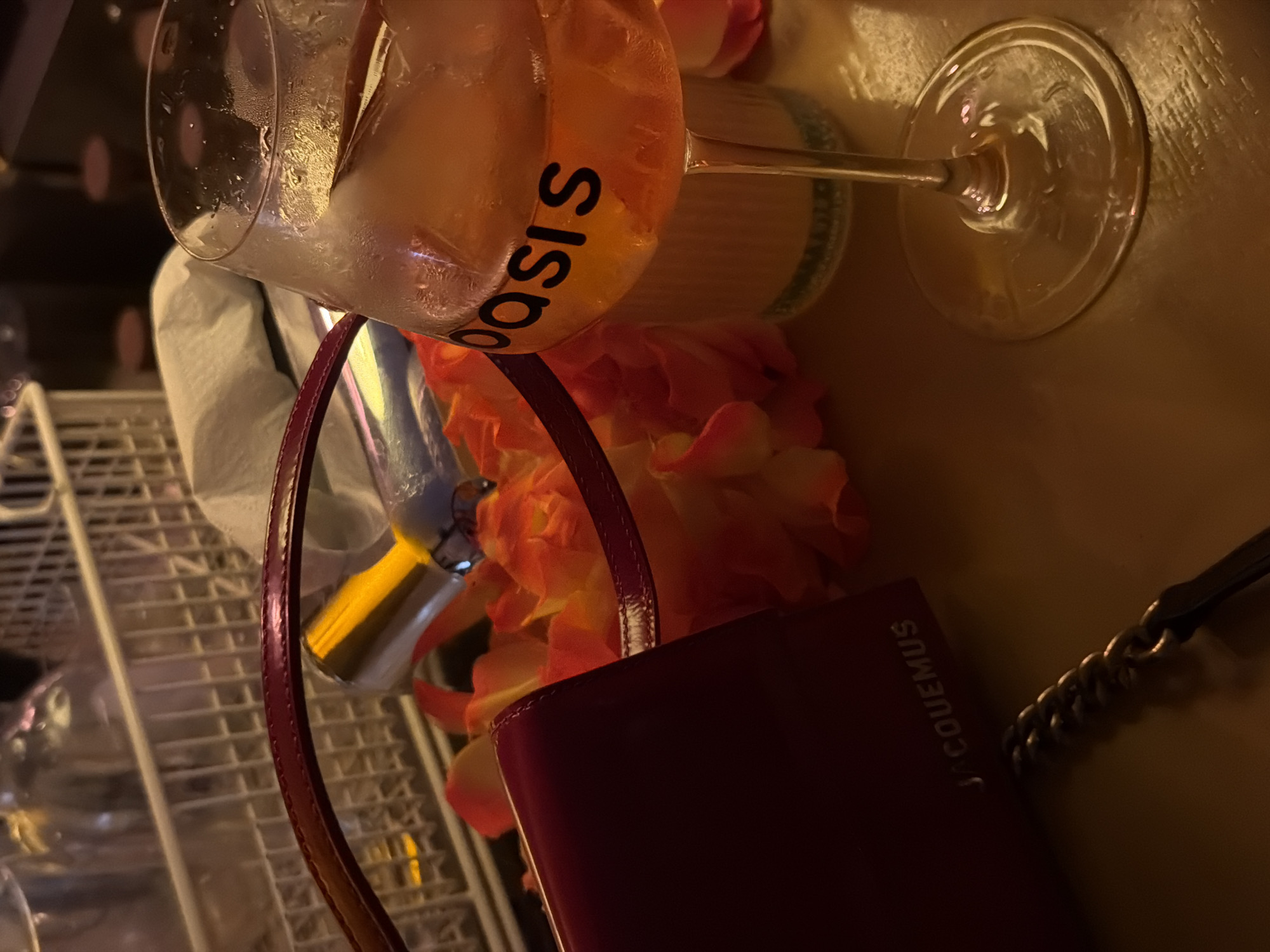 The image shows an overhead view of a table setting, likely in a bar or restaurant, as suggested by the presence of an "Oasis" branded cocktail glass filled with a pale orange drink, a wine glass, a Jacquemus handbag, a metal rack of glasses, a chain, flowers, and what appears to be a bar tool. The lighting is dim and warm-toned. There are no people visible in the image. The scene appears to be staged or posed. There is no way to verify if this image was taken in Krasnoyarsk, Russia.
